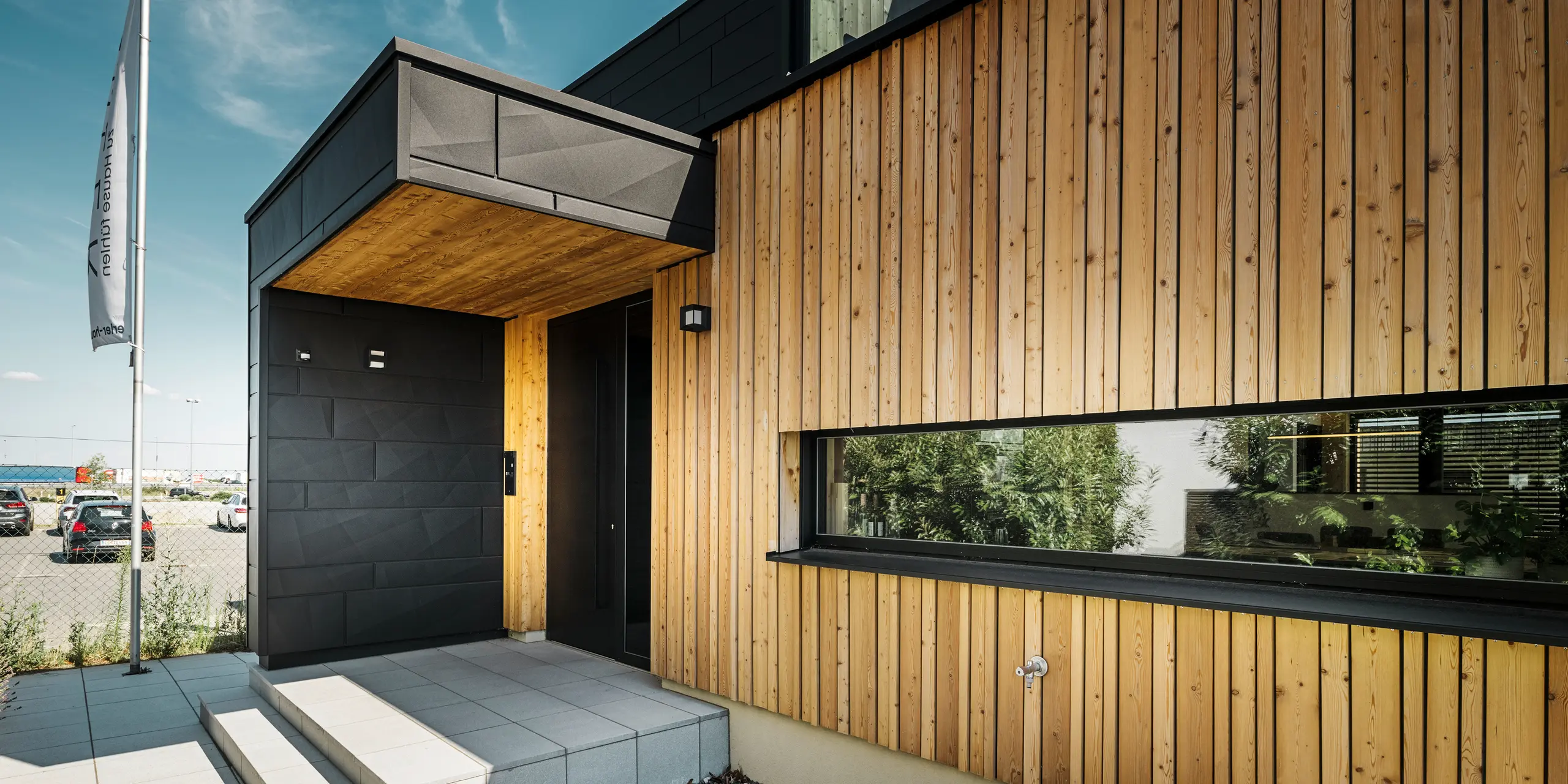Bočni ulaz u kuću Autark u centru montažnih kuća Blaue Lagune u Vösendorfu, s modernom fasadom od prirodnog drva i crnim PREFA Siding.X panelima. Slika prikazuje elegantnu kombinaciju drvene obloge i aluminija u visokokvalitetnom premazu P.10 crna.
