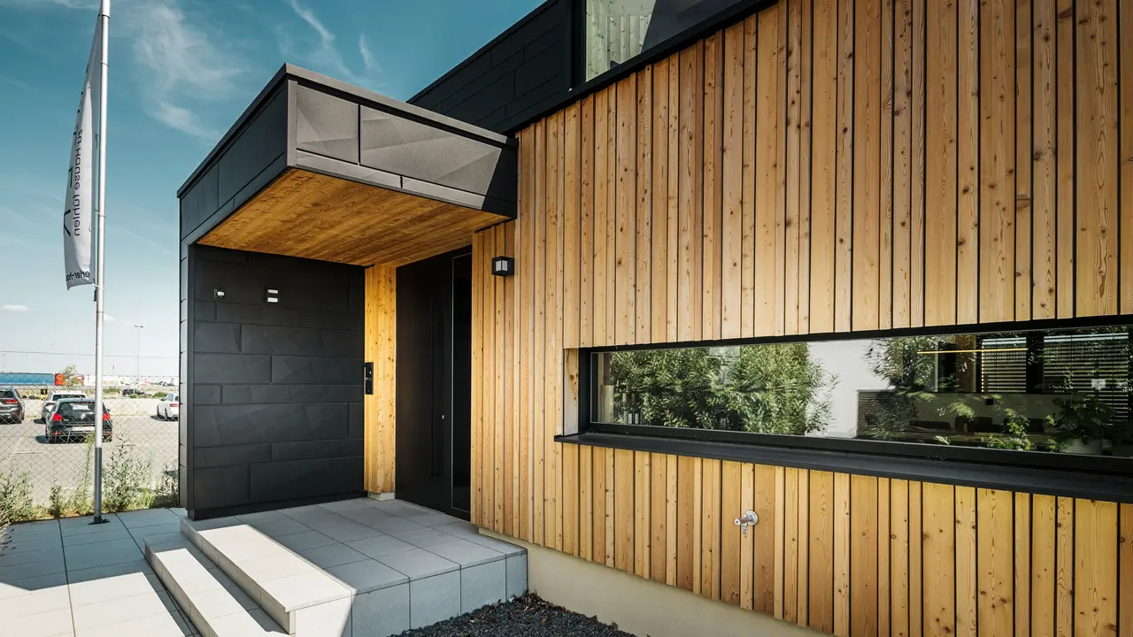 Bočni ulaz u kuću Autark u centru montažnih kuća Blaue Lagune u Vösendorfu, s modernom fasadom od prirodnog drva i crnim PREFA Siding.X panelima. Slika prikazuje elegantnu kombinaciju drvene obloge i aluminija u visokokvalitetnom premazu P.10 crna.
