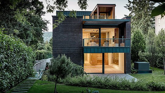 Stražnja strana moderne višegeneracijske vile u Budimpešti smještena je u bujnom zelenilu vrtnog krajolika. Velike staklene površine otvaraju stambeni prostor prema van i propuštaju toplo unutarnje svjetlo na strukturiranu fasadu od opeke. Jasne linije zgrade naglašene su PREFALZ aluminijem u boji P.10 antracit, koji kao obloga balkona i fasadni element daje dugotrajne akcentne detalje. Kombinacija opeke, aluminija i bogate vegetacije ističe bezvremenski, visokokvalitetan i arhitektonski zahtjevan karakter kuće.