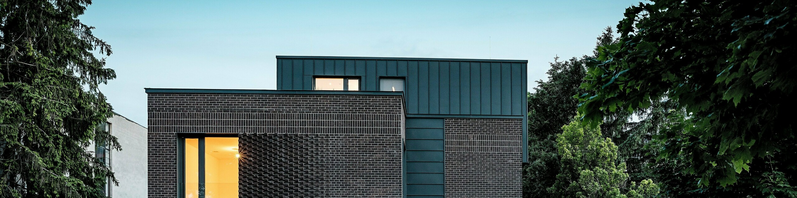 Snimka prikazuje modernu stambenu kuću u Budimpešti, čiji kubični krov od PREFALZ aluminija u boji P.10 antracit stvara upečatljiv kontrast prema povijesnom okruženju. Vertikalni stojeći falcevi stvaraju zanimljivu igru svjetla i naglašavaju jasni arhitektonski izraz. Visokokvalitetni PREFA aluminijski proizvodi daju zgradi ne samo elegantan i bezvremenski izgled, već se odlikuju i dugotrajnošću, otpornosti na vremenske uvjete i preciznom izradom. Antracitni dio fasade i krova predstavlja snažan vizualni naglasak i ističe inovativni karakter građevine.