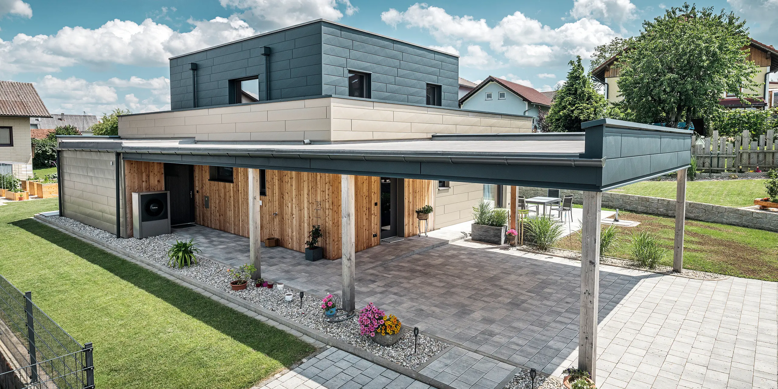 Pogled iz zraka na prilaz modernoj samostojnoj kući u Wippenhamu, Austrija, s dvobojnom aluminijskom fasadom PREFA u bronci i antracitu. Kuća prikazuje elegantnu integraciju fasadnih panela Siding.X i drvene obloge. Pokriveni natkriveni parking za automobile jasno je vidljiv i besprijekorno integriran u arhitekturu kuće. Uređeni vrt i terasni prostor naglašavaju moderan i funkcionalan dizajn zgrade.