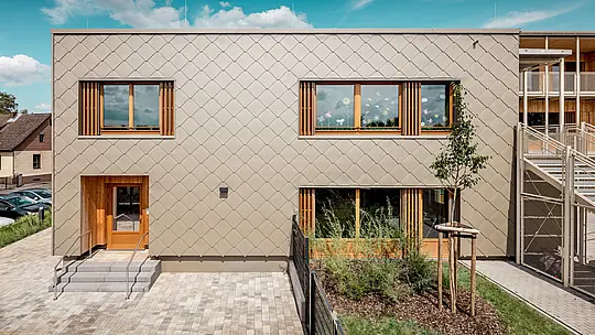 Stražnja strana moderne dječje ustanove u Alzenauu prikazana je iz frontalne perspektive i impresionira jasnim linijama te upečatljivom fasadom od brončano obojenih PREFA zidnih rombova 44 × 44. Geometrijska struktura fasade stvara skladan uzorak „ljuski“, koji je naglašen smirenim, horizontalnim rasporedom prozora. Prostrani drveni okviri i vertikalne lamele unose prirodne naglaske te stvaraju toplu i pozivajuću atmosferu. Promišljena kombinacija dugotrajnog aluminija i lokalnog drva simbolizira održivu arhitekturu visoke estetske kvalitete. Slika ističe preciznu izvedbu, vrhunski odabir materijala i uspješnu integraciju zgrade u urbani kontekst.