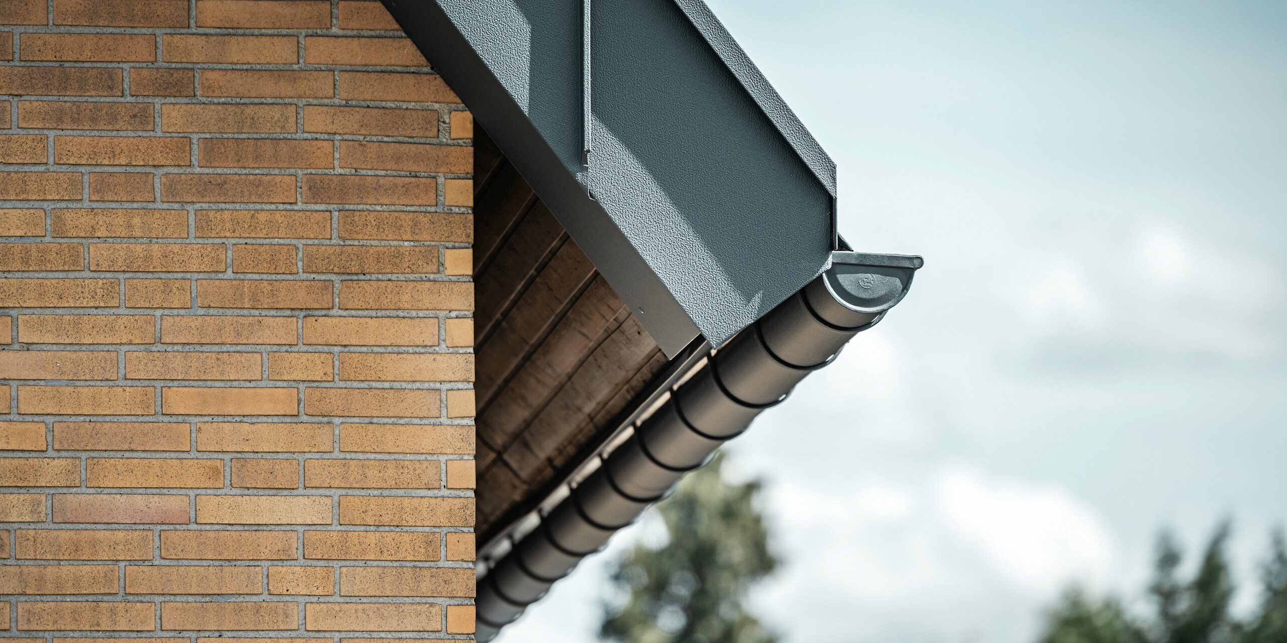 Detailaufnahme eines anthrazitfarbenen Dachentwässerungsystems von PREFA. Die Hängerinne hebt sich deutlich von den warmen Tönen der Backsteinwand ab und betont die präzise Verarbeitung und Qualität der verwendeten Materialien.