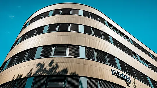 Dramatičan pogled ispod zakrivljenog novog dijela Policijske postaje 43 u Hamburgu otkriva upečatljivu PREFA aluminijsku fasadu od eloksiranih ekstrudiranih zupčastih profila u srednje brončanoj nijansi. Fino rebrasta površina stvara dinamične refleksije svjetla te dodatno ističe zaobljene linije objekta i horizontalne prozorske nizove. Zahvaljujući izvrsnoj otpornosti na koroziju, dugom vijeku trajanja i minimalnom održavanju, PREFA aluminijska rješenja omogućuju održivo, funkcionalno i estetski sofisticirano oblikovanje fasada.