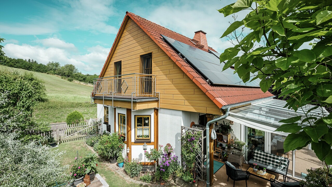 Ova slika prikazuje šarmantnu obiteljsku kuću u mjestu Blieskastel, čiji svježe obnovljen zabat s PREFA fasadnim kazetama u izvedbi prirodni hrast odmah privlači pozornost. Dugovječni aluminij u toplom izgledu drva upečatljivo je usklađen s crvenim ciglenim krovom i prostranim zimskim vrtom. Raskošno zelenilo i pažljivo raspoređene gredice s cvijećem doprinose ovom idiličnom, živahnom pejzažu. Robusna sanacija fasade uz proizvode tvrtke PREFA kući pruža prirodan izgled, ali i oduševljava svojom otpornošću na vremenske uvjete, postojanošću uvjerljivih boja i dugovječnošću koja zahtijeva minimalno održavanje.