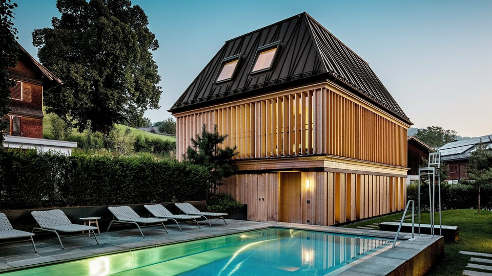 „Badehaus“ hotela Hirschen u Schwarzenbergu u sumrak, s pogledom na osvijetljeni vanjski bazen u prvom planu. Zgrada impresionira svojim modernim krovištem s četiri vode od PREFALZ-a u boji P.10 orah smeđa, s precizno izvedenim, dijelom dijagonalno postavljenim falcima. Dva krovna prozora osiguravaju dnevno svjetlo unutra. Fasada se sastoji od vertikalnih, etažom visokih drvenih lamela koje zgradi daju strukturu i lakoću. U donjem dijelu dominira prirodno drvo. Okružena njegovanim zelenilom i smještena u brežuljkastom krajoliku Bregenzerwalda, stvara se harmonična povezanost arhitekture, prirode i odmora.