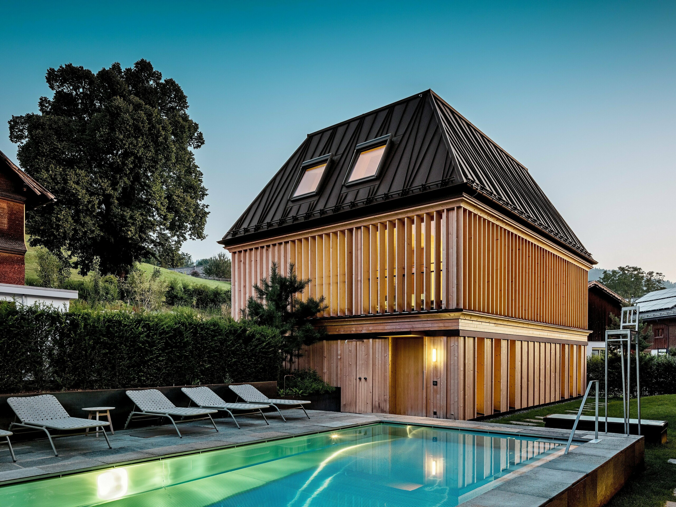 „Badehaus“ hotela Hirschen u Schwarzenbergu u sumrak, s pogledom na osvijetljeni vanjski bazen u prvom planu. Zgrada impresionira svojim modernim krovištem s četiri vode od PREFALZ-a u boji P.10 orah smeđa, s precizno izvedenim, dijelom dijagonalno postavljenim falcima. Dva krovna prozora osiguravaju dnevno svjetlo unutra. Fasada se sastoji od vertikalnih, etažom visokih drvenih lamela koje zgradi daju strukturu i lakoću. U donjem dijelu dominira prirodno drvo. Okružena njegovanim zelenilom i smještena u brežuljkastom krajoliku Bregenzerwalda, stvara se harmonična povezanost arhitekture, prirode i odmora.