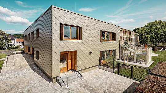 Slika prikazuje gradsku dječju ustanovu u Alzenauu iz dijagonalne ptičje perspektive, ističući modernu arhitekturu s upečatljivom PREFA aluminijskom fasadom u brončanoj boji. Fasada se sastoji od zidnih rombova 44 × 44, koji stvaraju skladan uzorak „ljuski“ i zgradi daju elegantnu dinamiku. Veliki drveni prozori s vertikalnim lamelama unose toplinu i propuštaju obilje dnevnog svjetla u unutrašnjost. S desne strane slike teren se otvara s ograđenim igralištem, zelenim površinama i natkrivenom terasom. Kombinacija dugotrajnog, bezdržavnog aluminija i održivih materijala čini zgradu uzorom funkcionalne i ekološki osviještene arhitekture u obrazovnom sektoru.