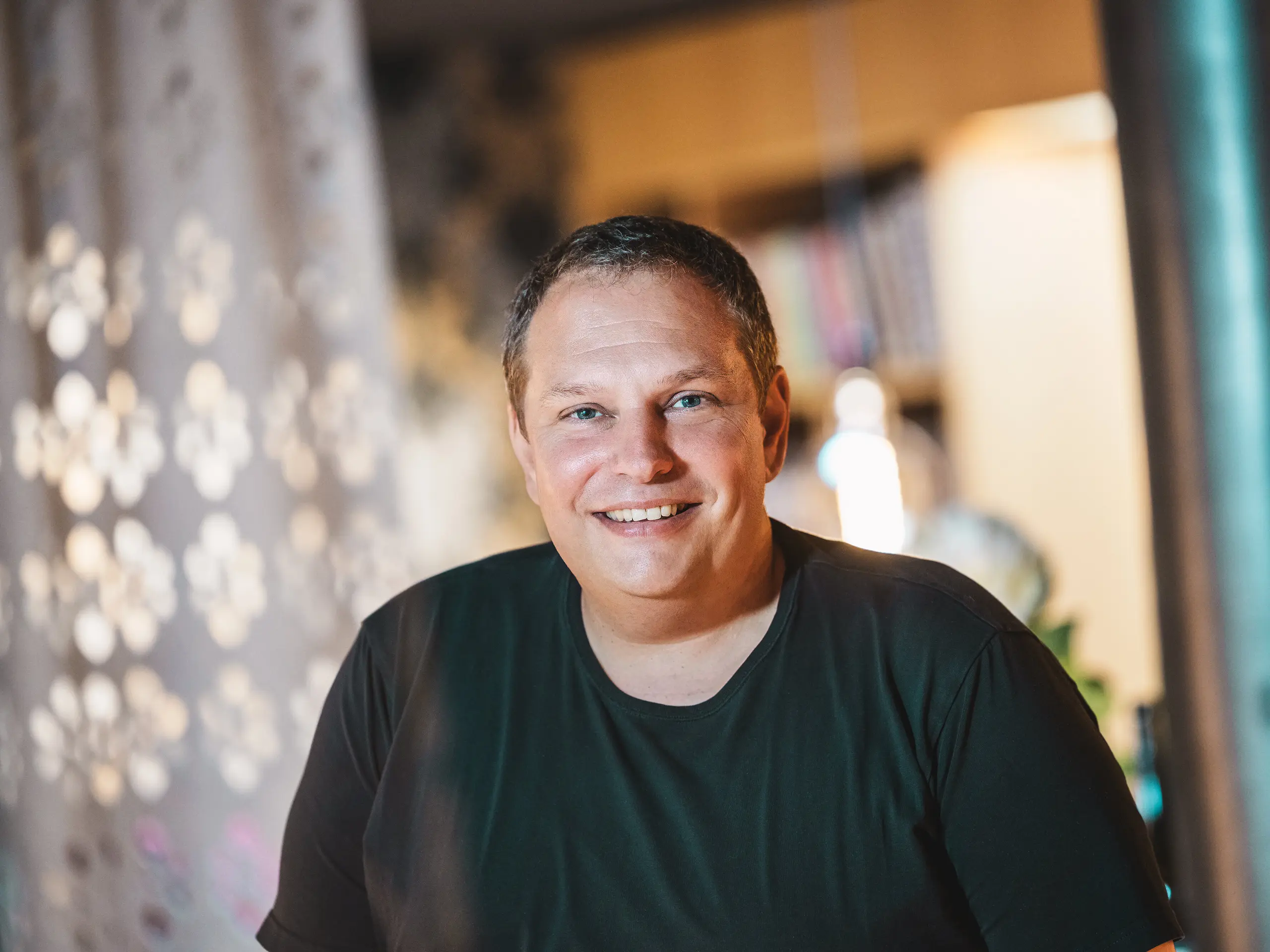 Portret vorarlberškog arhitekta Jürgena Hallera, koji je projektirao kuću Immi u Ebnitu kao dosljedno svedenu i održivu drvenu kuću. Nasmiješeni arhitekt stoji u toplinom osvijetljenom interijeru. Njegov projekt spaja regionalne materijale s robusnim PREFA solarnim krovom, koji se sastoji od solarnih krovnih ploča i krovnih ploča R.16 u boji P.10 Crna, prilagođenim zahtjevnim alpskim uvjetima.