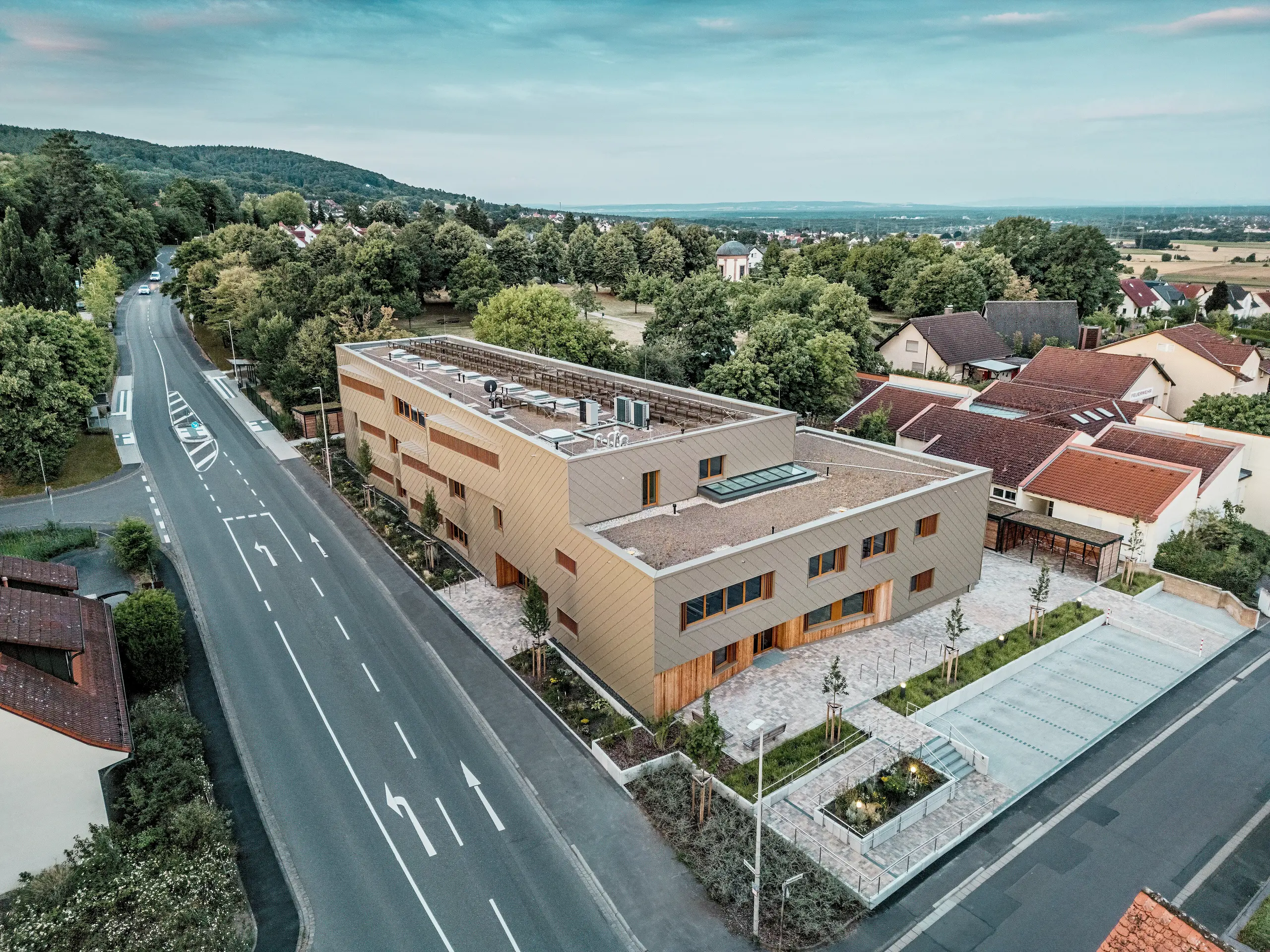 Impresivni ukupni dojam gradske dječje ustanove u Alzenauu postaje vidljiv iz zračne snimke. L‑oblik hibridne zgrade skladno je smješten u mirnu stambenu četvrt na rubu Spessarta. Fasada od brončano obojenih PREFA zidnih rombova 44 × 44 daje građevini upečatljiv, visokokvalitetan izgled i naglašava modernu arhitekturu. Zeleni krovovi, prostrani vanjski prostori i jasna organizacija prometa dodatno ističu održivi koncept cijelog projekta. Kombinacija dugotrajnog aluminija, drva i recikliranog betona zadovoljava najviše ekološke standarde. Ova perspektiva ističe promišljenu urbanističku integraciju i multifunkcionalni karakter zgrade – budućnosno orijentiran projekt za obrazovanje, stanovanje i održivost.