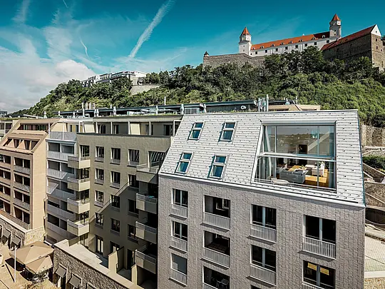 Moderna krovna arhitektura u gradskoj četvrti Vydrica u Bratislavi s pogledom prema kompleksu dvorca: strme krovne površine s velikim otvorima prozora i jasnim linijama elegantno naglašene su PREFA aluminijskim krovnim šindrama u bojama Prefa bijela i pješčano smeđa. Precizna izvedba i skladno povezivanje krova i fasade stvaraju snažan vizualni naglasak iznad krovova grada.  PREFA aluminij odlikuje se malom težinom, visokom otpornošću na vremenske uvjete i dugotrajnom kvalitetom – idealno rješenje za složene, arhitektonski zahtjevne stambene objekte.