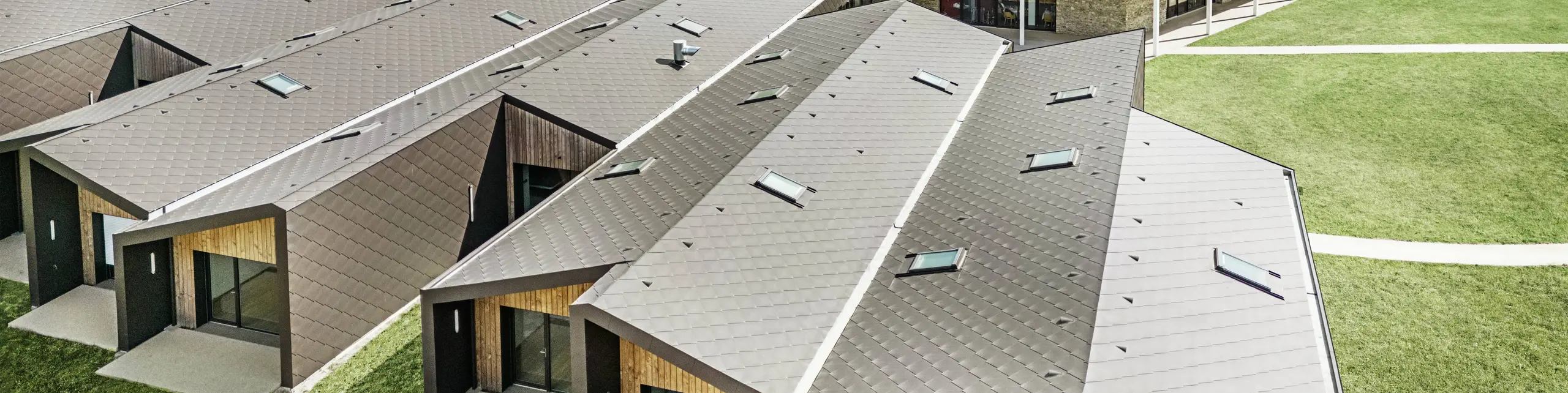 Slika prikazuje zračnu snimku modernog stambenog kompleksa, obloženog PREFA krovnim i zidnim rombovima 44 × 44 u orah smeđoj boji. Krovni pejzaž karakteriziraju geometrijski oblici, koji su kombinirani s velikim staklenim površinama i drvenim elementima. U pozadini se nalazi stara crkva, koja stvara skladan kontrast s modernom arhitekturom. Zeleni travnjak i okolna priroda dodatno naglašavaju otvoren i održiv dizajn cijelog kompleksa.