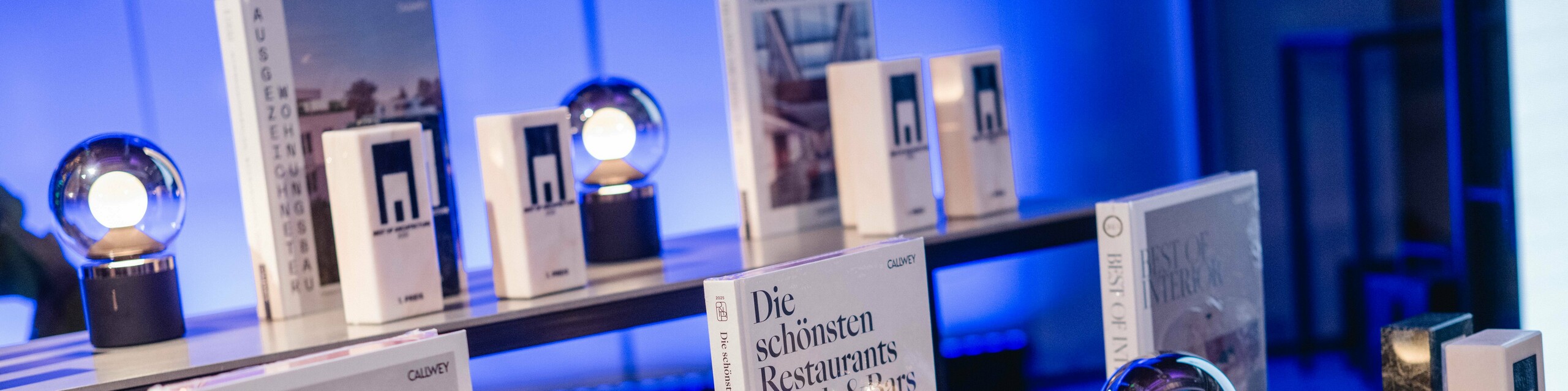 Izložba nagrada na natječaju „Stambene zgrade godine“ 2025.: nekoliko elegantnih trofeja s logom „Best of Architecture“ postavljeno je na moderan stol, uz visoko kvalitetne knjige o arhitekturi i dizajnu poput „Best Workspaces“ i „Die schönsten Restaurants Hotels & Bars“. Scena je obasjana atmosferičnim plavim svjetlom, što dočarava ekskluzivnost, inovaciju i arhitektonsku izvrsnost.