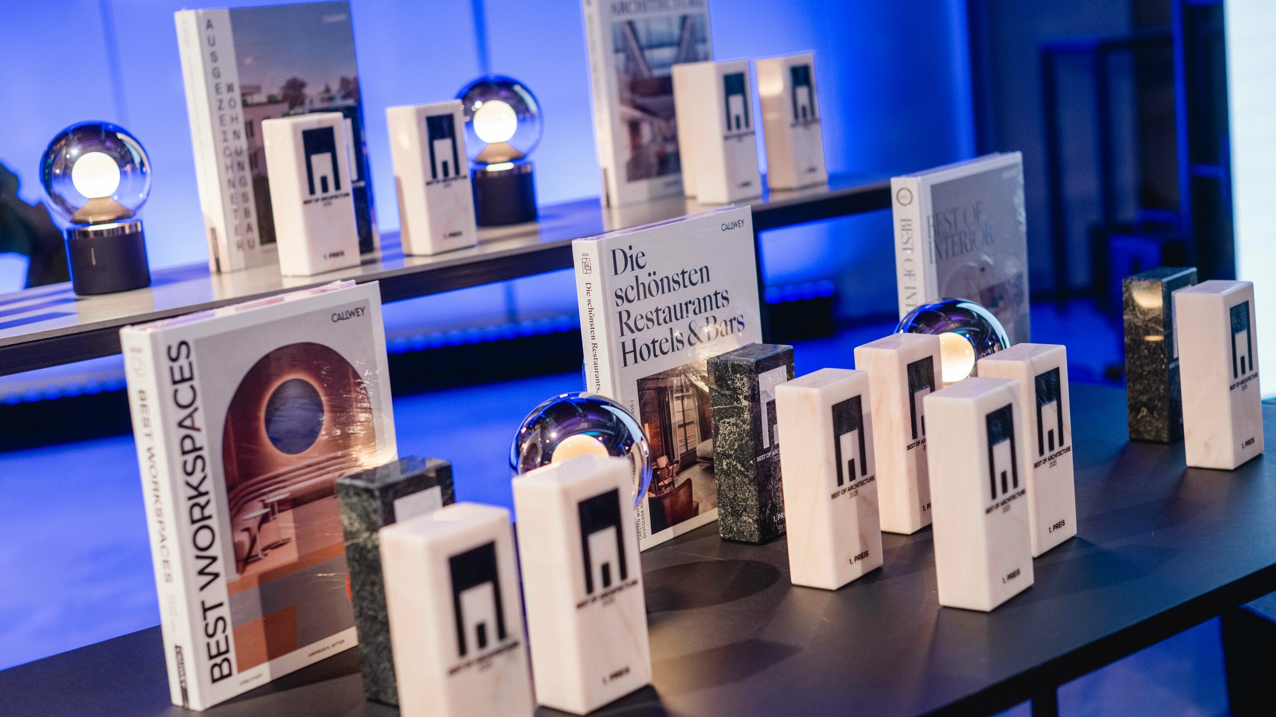 Izložba nagrada na natječaju „Stambene zgrade godine“ 2025.: nekoliko elegantnih trofeja s logom „Best of Architecture“ postavljeno je na moderan stol, uz visoko kvalitetne knjige o arhitekturi i dizajnu poput „Best Workspaces“ i „Die schönsten Restaurants Hotels & Bars“. Scena je obasjana atmosferičnim plavim svjetlom, što dočarava ekskluzivnost, inovaciju i arhitektonsku izvrsnost.