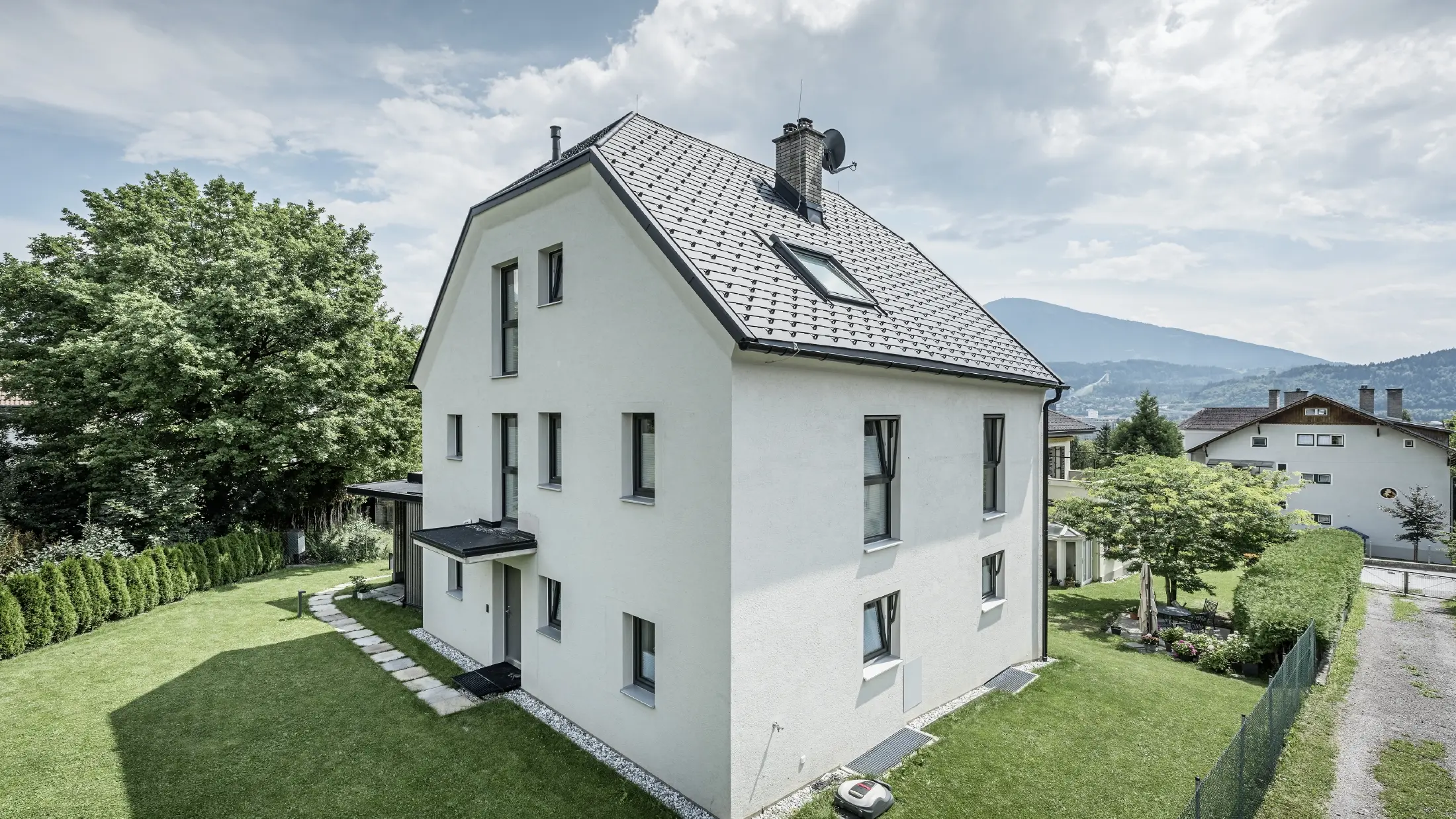 Moderna samostojeća kuća na tri kata s fasadom od svijetlog žbuka i tamnosivim aluminijskim krovom zabata iz PREFA. Zgrada ima nekoliko uskih prozora i krovna odvodnja u antracitnoj boji. Nalazi se u dobro održavanom vrtu s travnjakom, stablima i živicama, a u pozadini su vidljive druge stambene zgrade i planinski krajolik.
