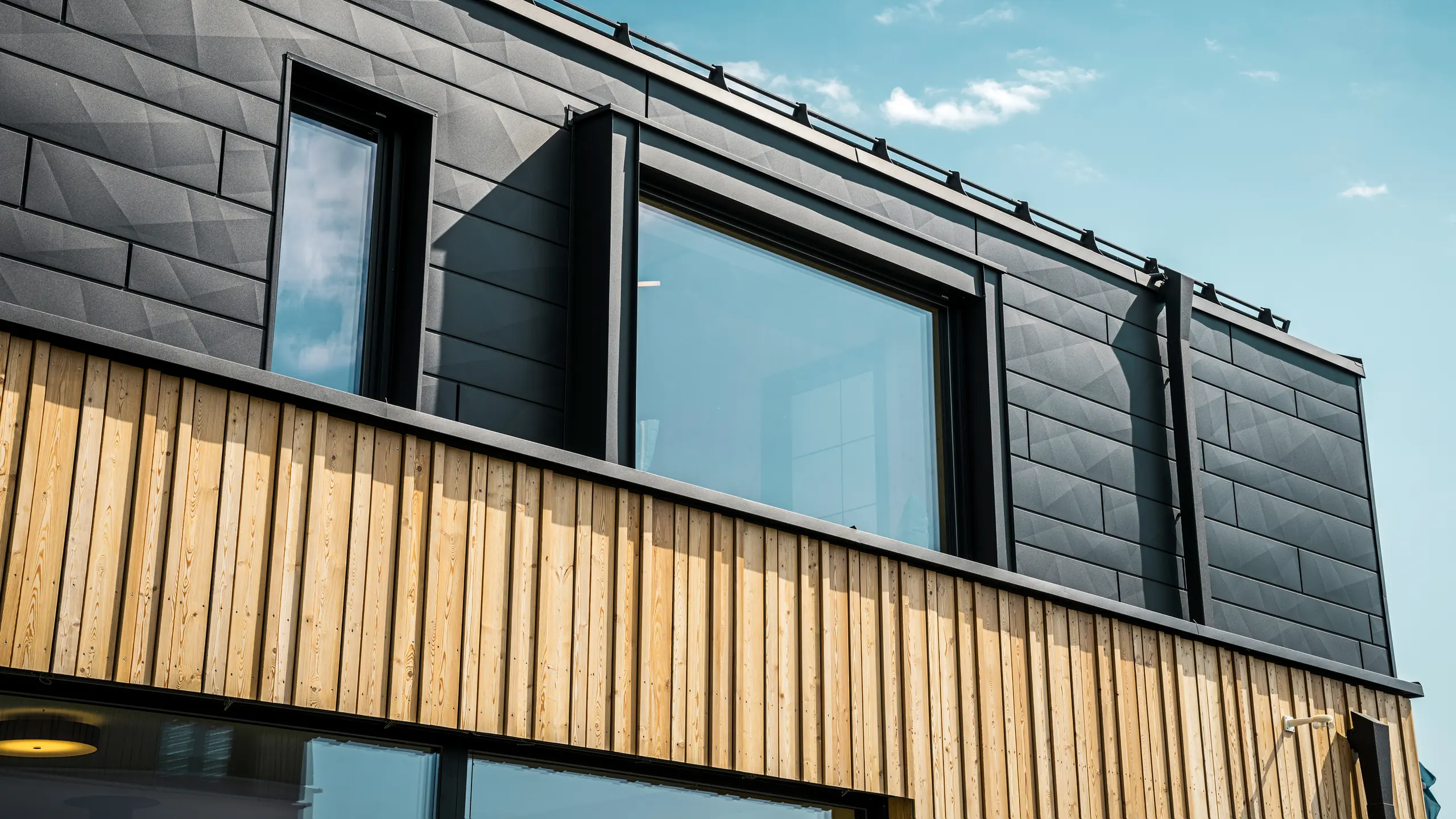 Krupni plan gornjeg kata modelne kuće Autark u centru montažnih kuća Blaue Lagune u Vösendorfu (AT). Fasada je izrađena od aluminijskih panela PREFA Siding.X u boji P.10 Black, koji oduševljavaju svojim prepoznatljivim izgledom s različitim rubovima. Ispod, elegantna drvena obloga stvara skladan kontrast s tamnim aluminijem. Veliki prozori propuštaju puno svjetla u unutrašnjost zgrade, naglašavajući moderan dizajn.