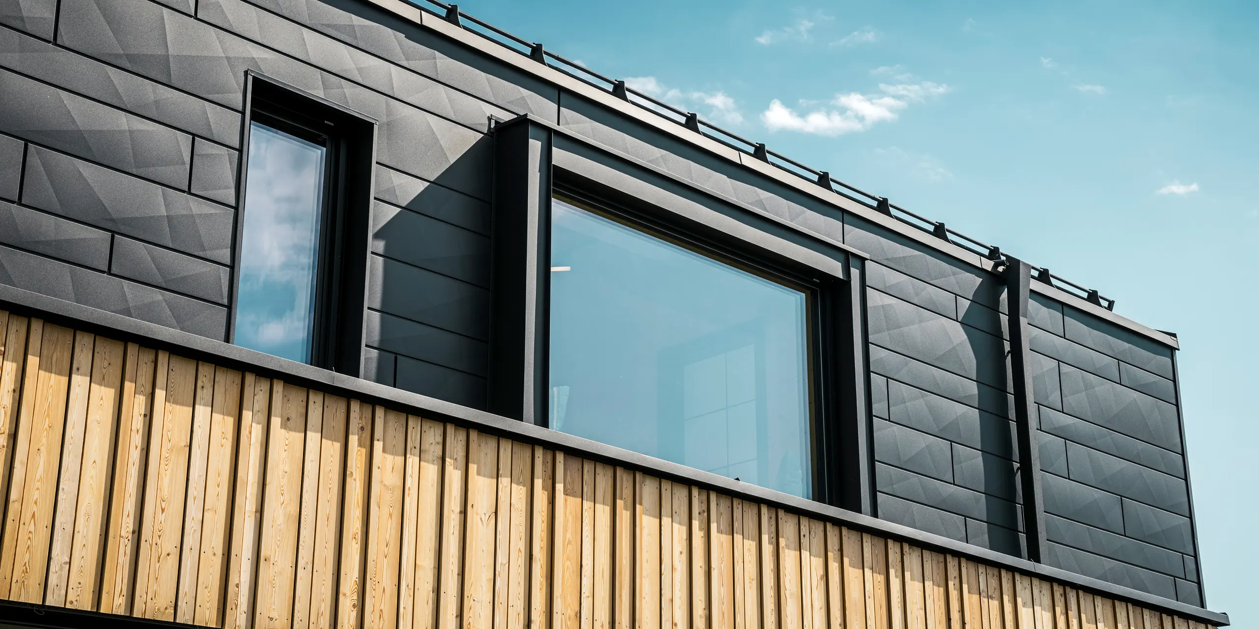 Krupni plan gornjeg kata modelne kuće Autark u centru montažnih kuća Blaue Lagune u Vösendorfu (AT). Fasada je izrađena od aluminijskih panela PREFA Siding.X u boji P.10 Black, koji oduševljavaju svojim prepoznatljivim izgledom s različitim rubovima. Ispod, elegantna drvena obloga stvara skladan kontrast s tamnim aluminijem. Veliki prozori propuštaju puno svjetla u unutrašnjost zgrade, naglašavajući moderan dizajn.