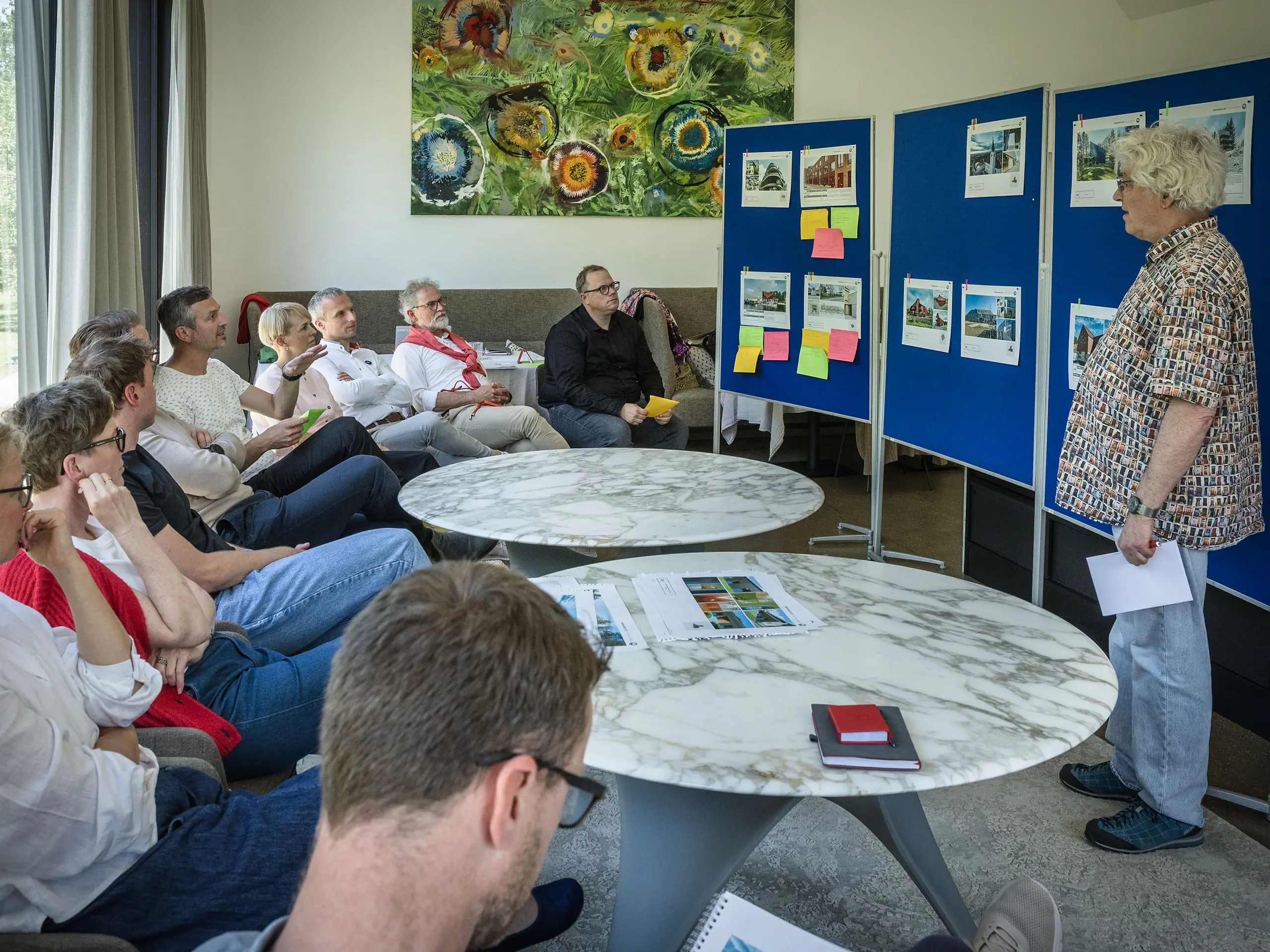 PREFARENZEN 2027 – sjednica žirija u svijetloj prostoriji za seminare: Moderator Matthias Finkentey predstavlja arhitektonske projekte od PREFA aluminija ispred plavih panoa, dok žiri pažljivo sjedi i bilježi dojmove. Veliki prozori propuštaju dnevno svjetlo i pružaju pogled na zeleni vrt. Na stolovima se nalaze arhitektonske knjige, bilježnice i olovke. Scena prenosi dojam usredotočene analize, kreativne razmjene ideja te vrednovanja dizajna i funkcionalnosti. PREFA aluminijski proizvodi simboliziraju dugotrajne, inovativne krovove i fasade koji objedinjuju estetiku, kvalitetu i inspiraciju za arhitektonske projekte.