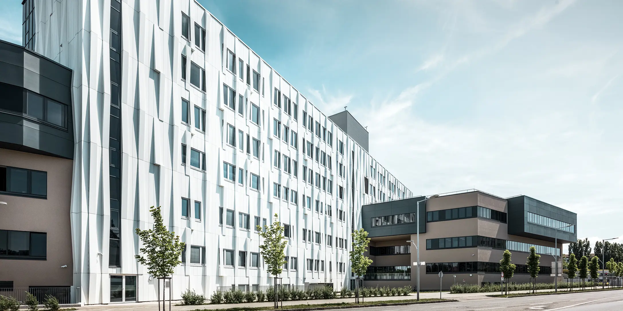Bočni pogled na Sveučilišnu bolnicu u St. Pöltenu, Donja Austrija. Robusna aluminijska fasada čini zgradu upečatljivim elementom u gradu. U prvom planu vidi se ulica s nekoliko stabala.
