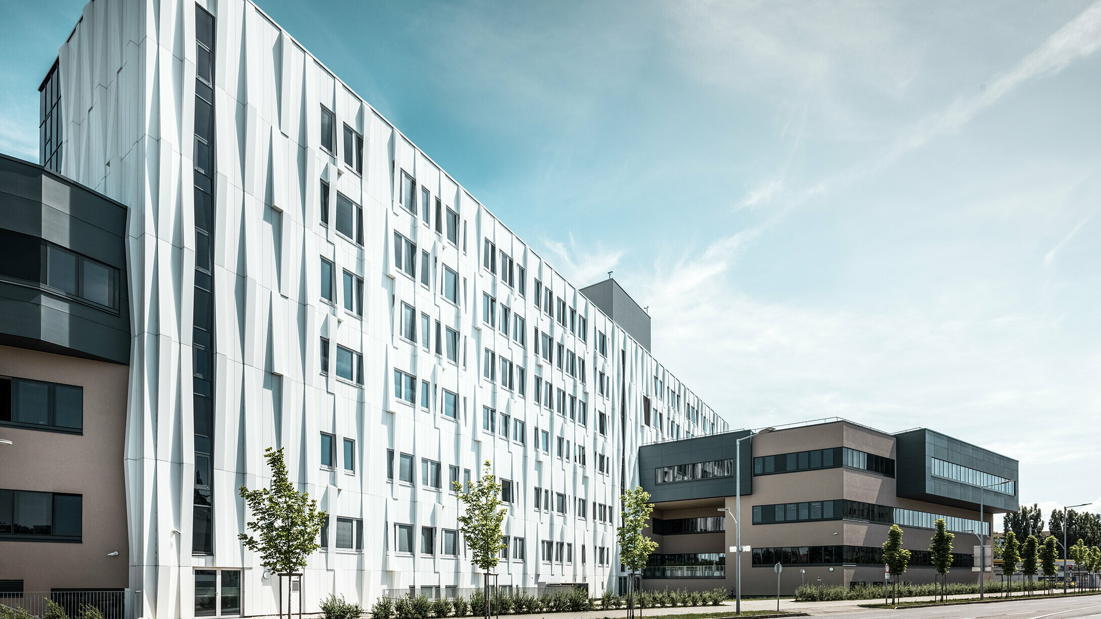 Bočni pogled na Sveučilišnu bolnicu u St. Pöltenu, Donja Austrija. Robusna aluminijska fasada čini zgradu upečatljivim elementom u gradu. U prvom planu vidi se ulica s nekoliko stabala.