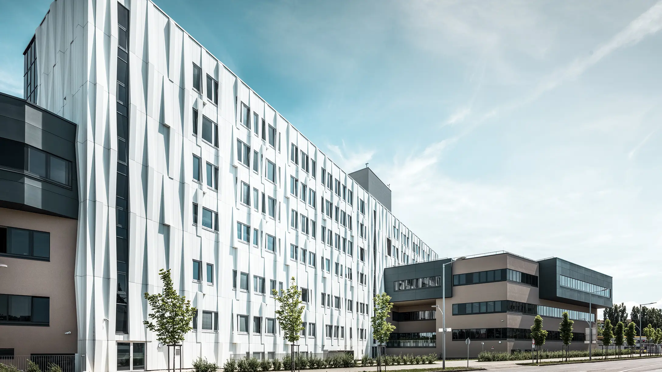 Bočni pogled na Sveučilišnu bolnicu u St. Pöltenu, Donja Austrija. Robusna aluminijska fasada čini zgradu upečatljivim elementom u gradu. U prvom planu vidi se ulica s nekoliko stabala.