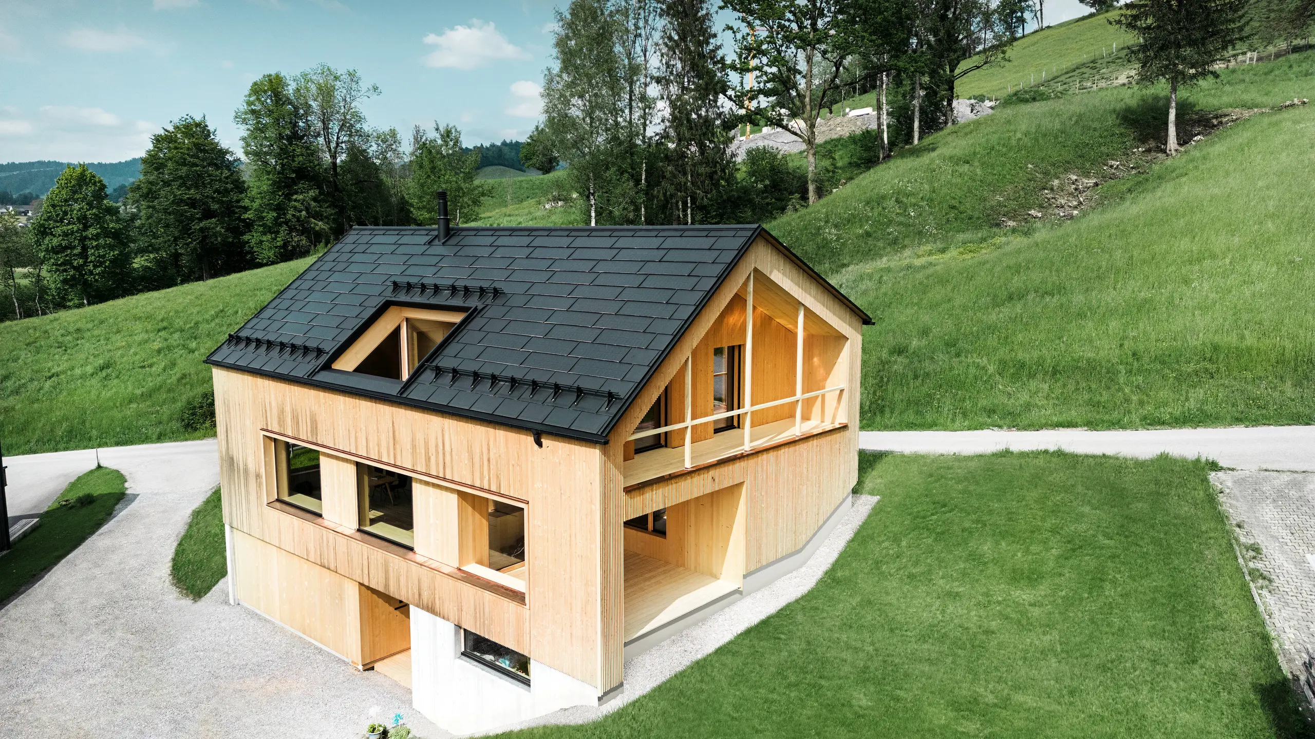 Slika prikazuje slikovitu ekološku kuću u Eggu, smještenu među valovitim zelenim brežuljcima, s održivim PREFA solarnim krovom. Crni, visokokvalitetni PREFA krovna ploča R.16 i PREFA solarna krovna ploča skladno se uklapaju u ruralni krajolik i naglašavaju prirodnu ljepotu okoliša. Solarna krovna ploča nisu samo posvećenost obnovljivoj energiji, već i dizajnerski element koji elegantno nadopunjuje modernu drvenu fasadu i čiste linije zgrade. Ova kuća je primjer ekološki osviještene gradnje i inspiracija za arhitekturu usmjerenu na budućnost.