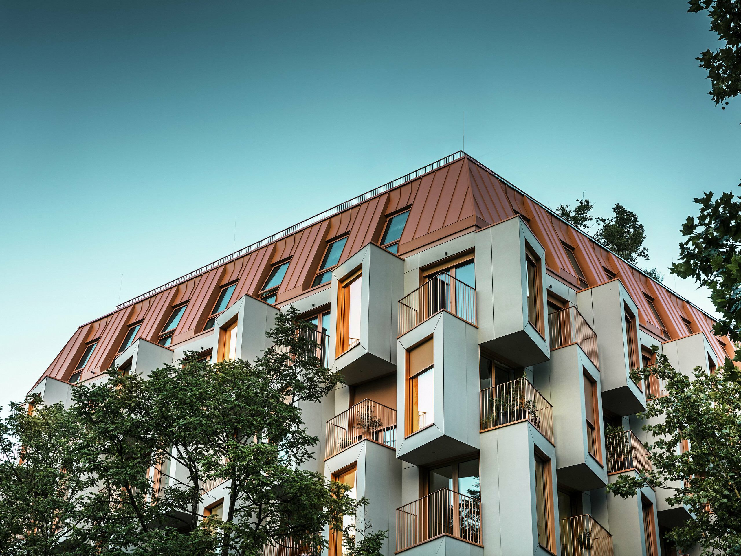 Arhitektonski istaknuta fasada stambenog projekta Van B u Münchenu s upečatljivim erkerima i PREFALZ aluminijskim krovom u posebnoj boji IGP Orange HDP. U sumrak pogled odozdo prikazuje svjetla u prozorima. Moderno krovno rješenje spaja vrhunsku otpornost na vremenske uvjete s elegantnim izgledom i ističe se bojom u gradskom prostoru. Igra između betona ojačanog staklenim vlaknima, bakreno-narančastih okvira prozora i aluminijskog krova stvara dinamičnu, dugotrajnu i održivu ovojnicu zgrade – idealno za budućnosti orijentiranu gradnju.