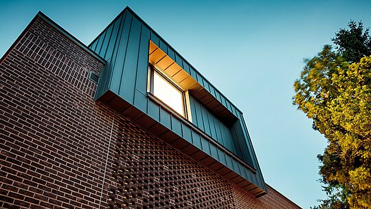 Die Aufnahme zeigt die moderne Fassadengestaltung der Budapester Mehrgenerationen‑Villa aus einer dynamischen Froschperspektive. Der auskragende Kubus mit PREFALZ Aluminium in P.10 Anthrazit bildet einen markanten Kontrast zu den dunkelroten Backsteinen darunter. Die präzise Ausführung der vertikalen Winkelstehfalze betont die klare Linienführung und unterstreicht die hohe Qualität der PREFA Aluminiumprodukte. Durch das warme Licht entsteht ein kraftvolles Zusammenspiel aus Metall, Klinker und Schatten, das den innovativen, zeitlosen Charakter des Gebäudes eindrucksvoll hervorhebt.