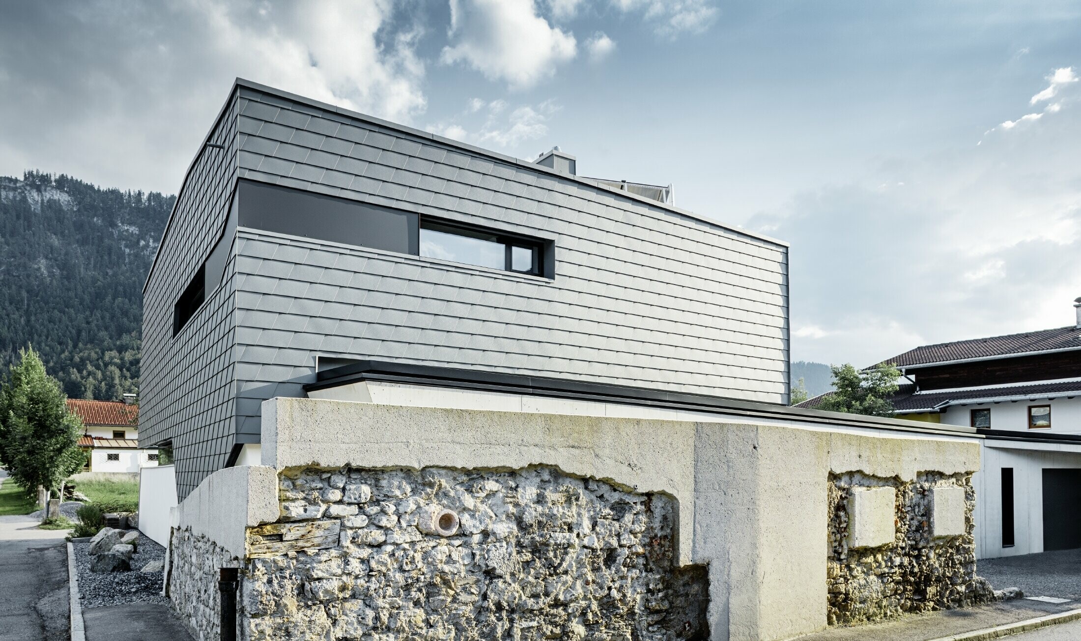 Moderna obiteljska kuća s ravnim krovom, velikim površinama prozora i fasadom iz šindre u svijetlo sivoj boji iz aluminija od PREFE