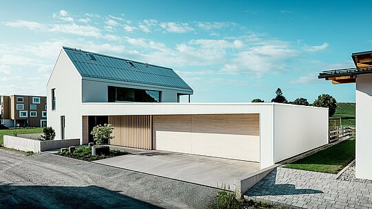 Slika prikazuje modernu obiteljsku kuću u Freistadtu, koja se ističe svojom jednostavnom, geometrijskom formom i kombinacijom čistobijele PREFALZ-aluminijske fasade i drvenih obloga. Drvena obloga na garažnim vratima pruža topao, prirodan kontrast i savršeno se uklapa u moderno dizajnirano rješenje. Krov, također prekriven PREFALZ-om, osigurava besprijekornu, otpornu na vremenske uvjete površinu. Ova kombinacija aluminija koji zahtijeva malo održavanja i drva stvara estetski privlačno i funkcionalno rješenje za održivu arhitekturu. Prostrano zeleno dvorište i vedro nebo u pozadini naglašavaju modernu kvalitetu života koju ova kuća pruža.