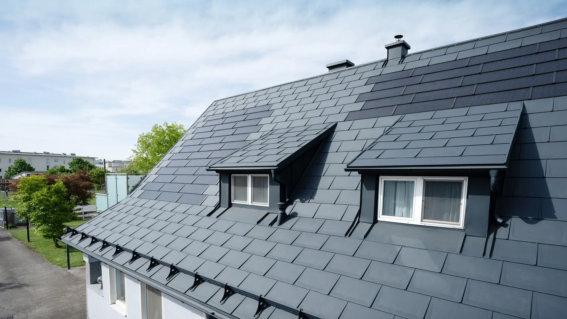 Krupni plan krova prekrivenog velikim PREFA solarnim krovnim panelima i PREFA R.16 krovnim panelom u boji P.10 Antracit. Vidljiva su dva šiltera. Solarni krovni paneli ravnomjerno su raspoređeni po krovu, pružajući moderno, održivo rješenje.