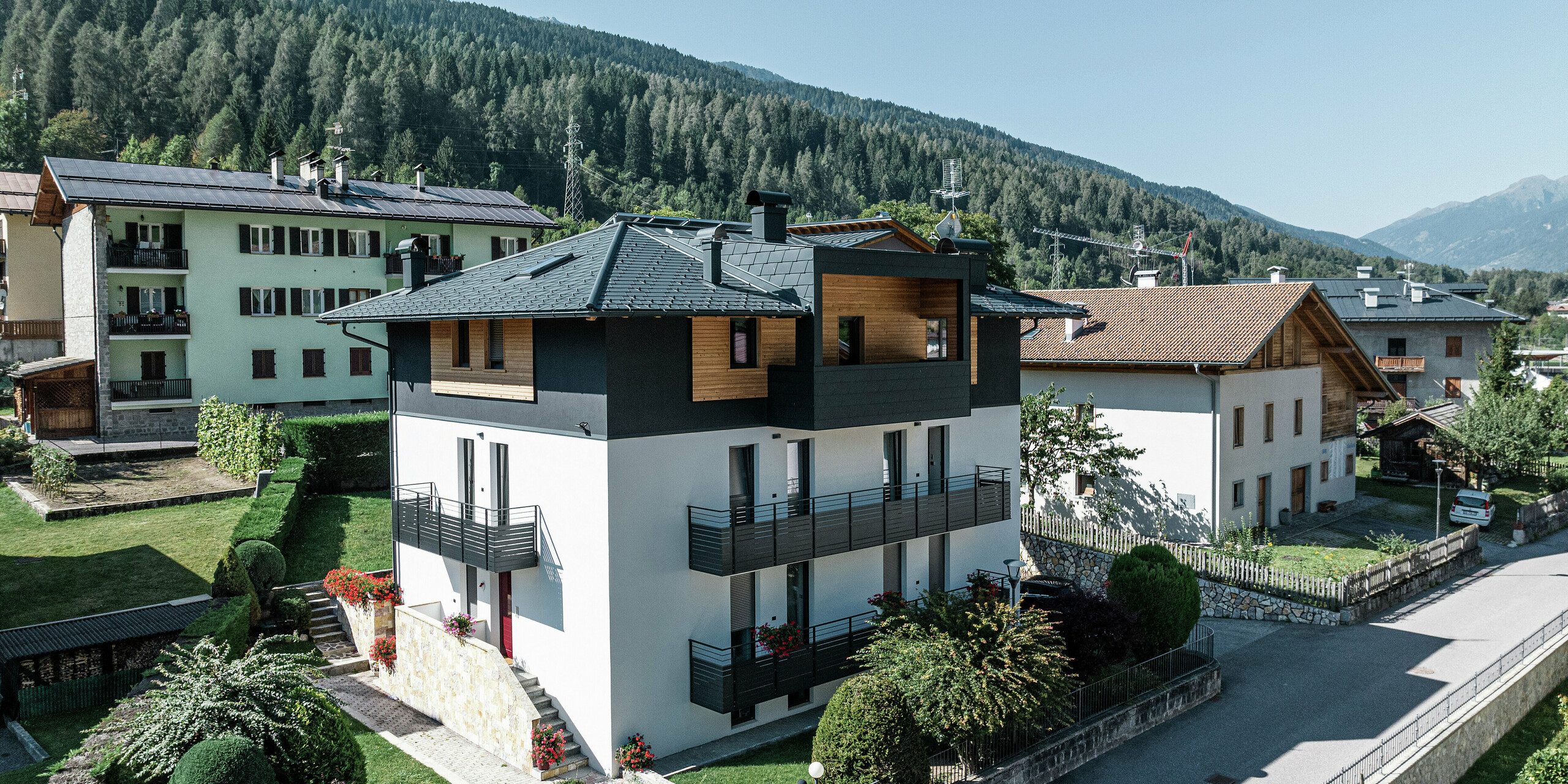Vorderseite des Wohnhauses in Commezzadura mit PREFA Dachschindeln DS.19 in P.10 Anthrazit. Die hochwertigen Aluminiumschindeln schützen sowohl das Dach als auch die Fassade der überdachten Balkone. Einzelne Holzelemente an der Fassade bilden einen gelungenen Übergang zur robusten Aluminiumverkleidung. Die Architektur harmoniert perfekt mit der alpinen Umgebung.