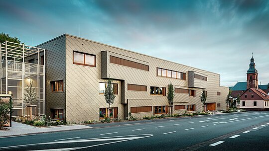 Snimka s razine ulice prikazuje novu dječju ustanovu u Alzenauu u dinamičnom dijalogu s povijesnom gradskom kulisom, uključujući upečatljivu crkvu u pozadini. Moderna fasada od brončano obojenih PREFA zidnih rombova 44 × 44 daje zgradi elegantan, gotovo skulpturalan dojam. Precizno postavljeni prozori, vertikalne drvene lamele i pomaknuti fasadni elementi stvaraju dubinu i ritam. S lijeve strane vidljivo je filigransko stubište s bijelom metalnom konstrukcijom koje zrači modernom lakoćom. Jasna oblikovna koncepcija, održiv odabir materijala i visokokvalitetna aluminijska fasada čine ovu zgradu primjerom uspješne urbane integracije i budućnosno orijentirane gradnje s PREFA sustavima.