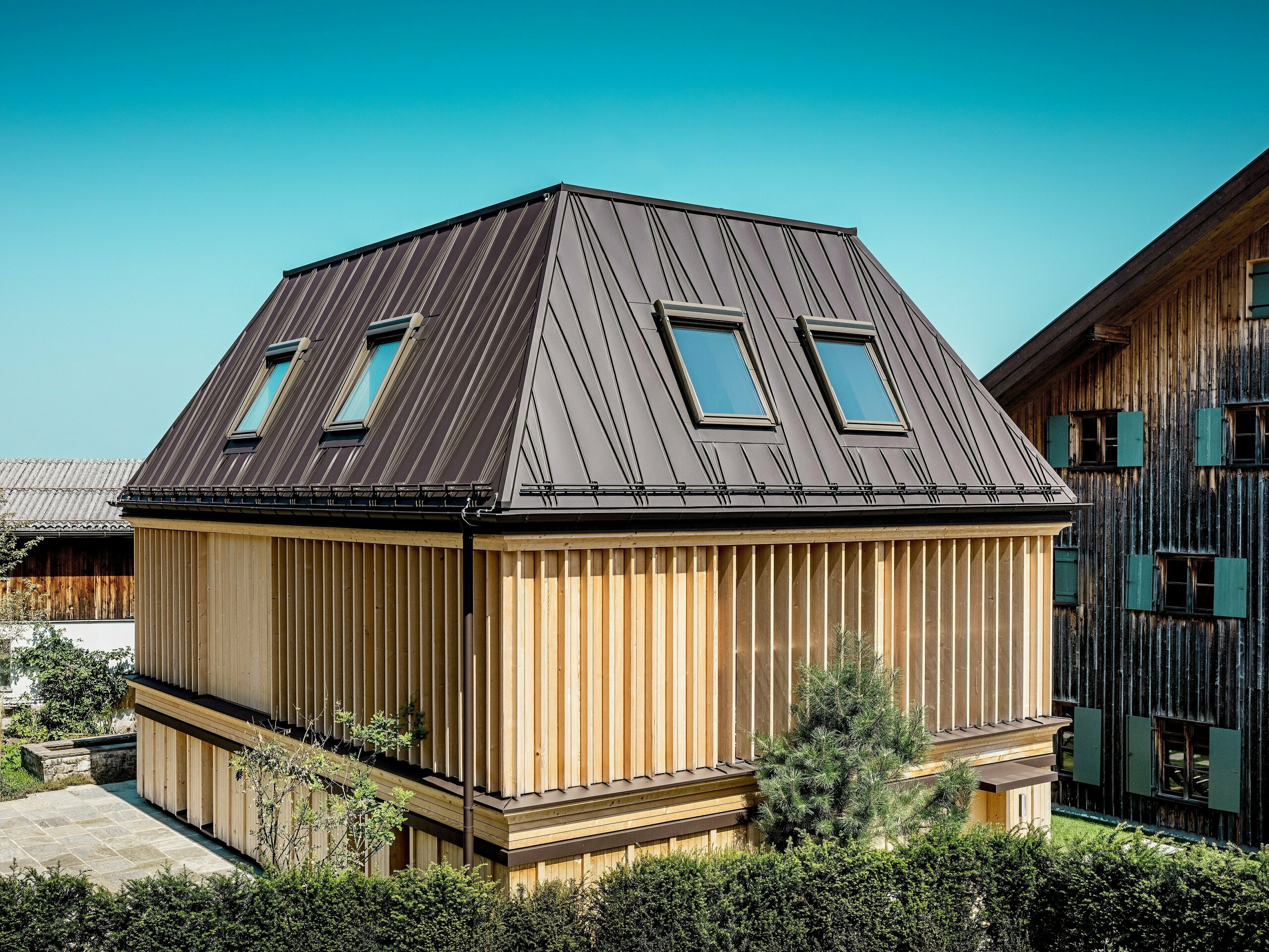 Novouređeni „Badehaus“ hotela Hirschen u Schwarzenbergu na dnevnom svjetlu. U prvom planu ističe se upečatljiv PREFA krov s falcom u boji P.10 orah smeđa, čiji atipično postavljeni falci krovu s četiri vode daju dinamičan, moderan izgled. Nagib krova od 60 stupnjeva naglašava izraženu arhitekturu. Fasada se sastoji od vertikalno postavljenih, etažom visokih drvenih lamela koje efektno upravljaju svjetlom i sjenom, a istovremeno pružaju funkcionalnu zaštitu od sunca. Povezanost tradicionalnog oblika gradnje i inovativnog izbora materijala stvara zanimljiv kontrast prema zaštićenoj postojeće zgradi u pozadini, čija drvena, izblijedjela fasada tipična je za regiju Bregenzerwald.