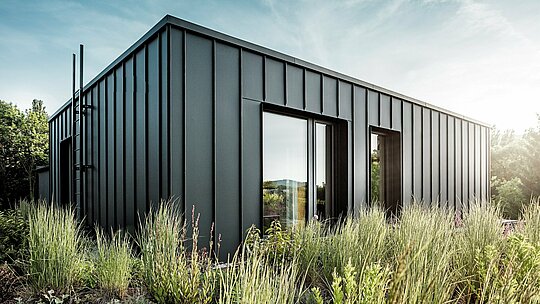 Snimka prikazuje modernu, kubičnu krovnu terasu budimpeštanske višegeneracijske vile, obloženu visokokvalitetnim PREFALZ aluminijem u boji P.10 antracit. Jasno strukturirani stojeći falcevi stvaraju elegantnu vertikalnu linijsku kompoziciju, koja je dodatno naglašena blagim sunčevim svjetlom. Okružena gustom, prirodnom vegetacijom, nastaje snažan kontrast između moderne metalne arhitekture i bujnog zelenila. Dugotrajni i vremenski otporni PREFA aluminijski proizvodi naglašavaju visokokvalitetnu izvedbu i pretvaraju krovnu terasu u upečatljiv arhitektonski element.