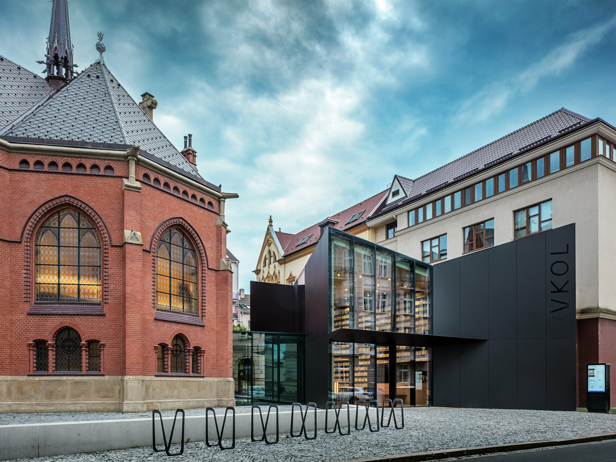 Slika prikazuje fascinantnu povezanost povijesne i suvremene arhitekture u Olomoucu. S lijeve strane stoji veličanstvena Crvena crkva sa svojim karakterističnim gotičkim prozorima i crvenim opečnim zidovima, dok se nova zgrada s desne strane ističe modernom fasadom od crnih PREFABOND aluminijsko-kompozitnih ploča. Staklena pročelja nove zgrade omogućuju pogled u inovativni koncept knjižnice i daju ansamblu elegantan, otvoren karakter. Precizna obrada PREFABOND ploča osigurava ujednačen izgled koji se skladno uklapa u povijesno okruženje, a istovremeno postavlja moderan vizualni naglasak. Arhitektura ovog projekta savršeno povezuje tradiciju i inovaciju, pri čemu materijali nisu samo estetski dojmljivi, već osiguravaju i dugovječnost te održivost.