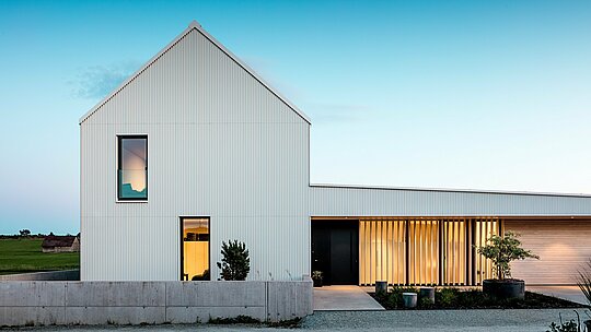 Slika prikazuje impresivnu obiteljsku kuću u Freistadtu, koja se ističe svojom upečatljivom, geometrijskom arhitekturom. Fasada zgrade izrađena je od precizno naboranog, čistobijelog PREFALZ-aluminija, što kući daje modernu eleganciju. Vertikalna struktura fasade i jasno, minimalističko oblikovanje naglašavaju visokokvalitetan odabir materijala i osiguravaju dugotrajnu, otpornu na vremenske uvjete površinu. Strmi zabatni krov, također prekriven PREFALZ-om, štiti od vremenskih utjecaja i istovremeno doprinosi skladnoj ukupnoj estetici. Jasna forma i korištenje aluminija omogućuju optimalno zadovoljenje estetskih i funkcionalnih zahtjeva. Prozori ispunjeni svjetlom i otvoreni ulaz djeluju pozivajuće i stvaraju zanimljiv kontrast prema modernoj fasadi. Odluka za PREFALZ naglašava arhitektonski standard i predstavlja održivo, lako održivo rješenje za dugotrajne građevinske projekte.