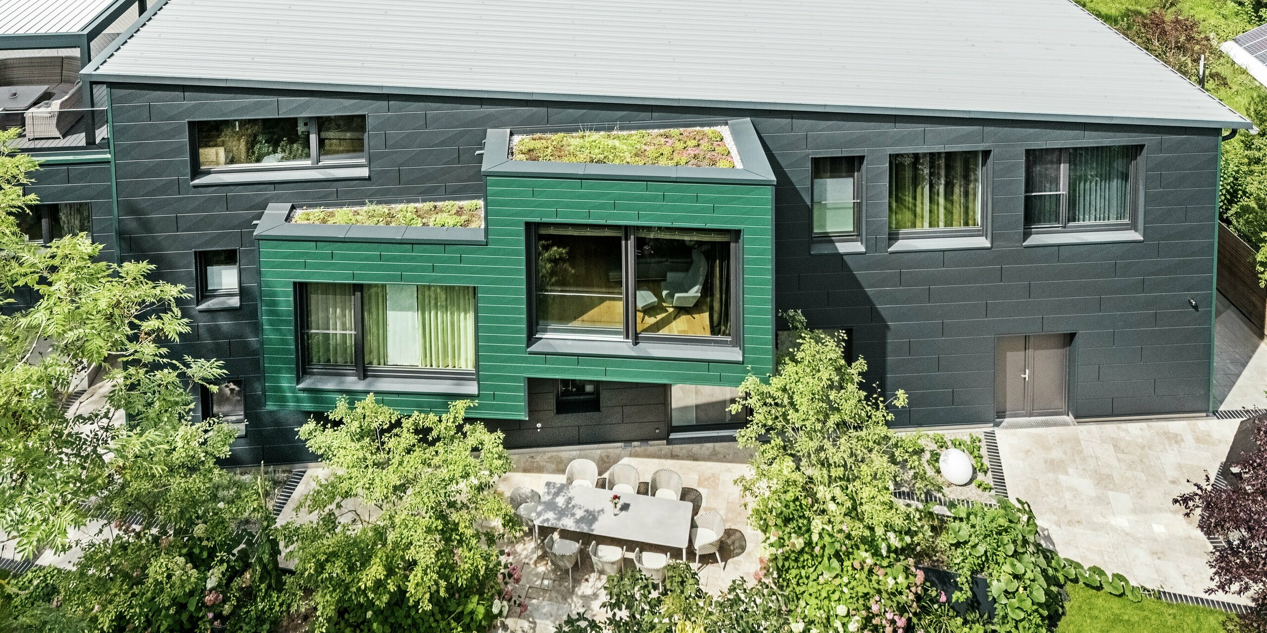 Dieses Luftbild zeigt das moderne Einfamilienhaus in Türkheim mit seiner beeindruckenden Fassade aus PREFA Siding X-Paneelen in den Farben Moosgrün und Anthrazit. Besonders auffällig ist das begrünte Flachdach, das dem Gebäude eine natürliche und nachhaltige Note verleiht. Im Vordergrund befindet sich eine elegante Terrasse mit stilvollen Gartenmöbeln und üppigen Pflanzen, die das harmonische Zusammenspiel von Architektur und Natur unterstreichen. Die pflegeleichte Aluminiumfassade schützt das Haus und sorgt für lang anhaltende, zeitlose Schönheit.