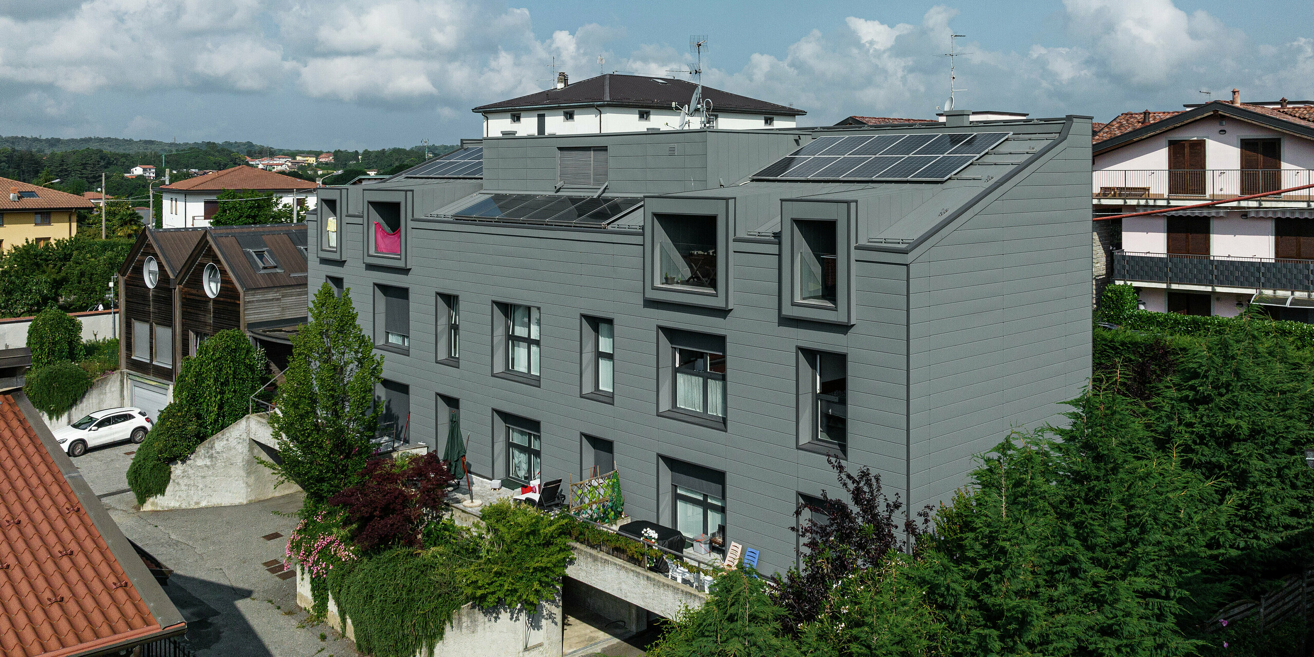 Weiträumiger Blick auf die moderne Wohnanlage in Faloppio, Italien, mit einer markanten Fassade aus PREFA Sidings und einem energieeffizienten Dach mit Photovoltaikmodulen. Diese Kombination aus innovativer Architektur und nachhaltiger Energiegewinnung ist ein Musterbeispiel für modernes und nachhaltiges Bauen. Die Blechfassade sorgt für Langlebigkeit und minimalen Wartungsaufwand, die Solarzellen für eine umweltfreundliche Stromquelle.