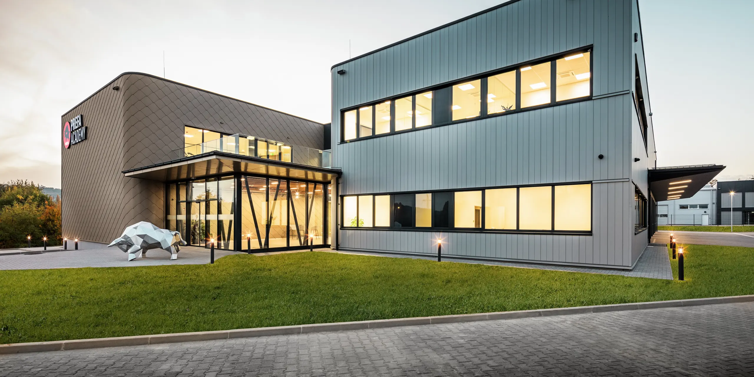 Der neue PREFA Standort in Törökbálint (HU) erstrahlt in der Abenddämmerung in modernem Design. Die Fassade kombiniert PREFA Wandrauten 44 × 44 in Bronze mit PREFA Sidings in Silbermetallic zu einer langlebigen und witterungsbeständigen Gebäudehülle. Großzügige Fensterflächen sorgen für eine helle und freundliche Atmosphäre, die ikonische Stierskulptur symbolisiert die Stärke der Marke PREFA.