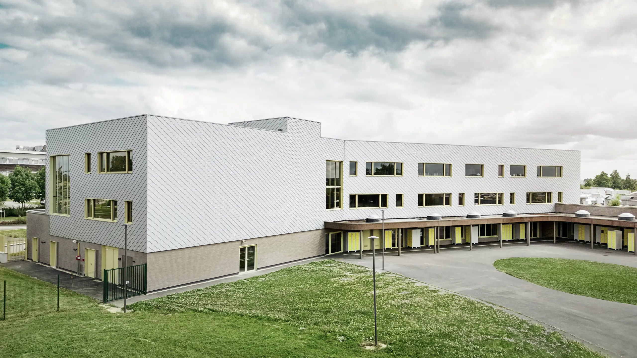 Slika prikazuje stražnju stranu Collège Léontine Dolivet. U prvom planu nalazi se prostrano asfaltirano školsko dvorište s jasnim linijama i otvorenim površinama, pogodnim za odmore i slobodne aktivnosti. Nekoliko zelenih površina razbija monotoniju okoline. U pozadini se uzdiže moderna školska zgrada s upečatljivom aluminijskom fasadom od srebrno-metalnih PREFA zidnih rombova, koja reflektira svjetlo i stvara dinamičan efekt. Više velikih prozora osigurava svjetlost u unutrašnjosti. Scena je snimljena danju, s lagano oblačnim nebom, što posebno naglašava metalnu površinu.