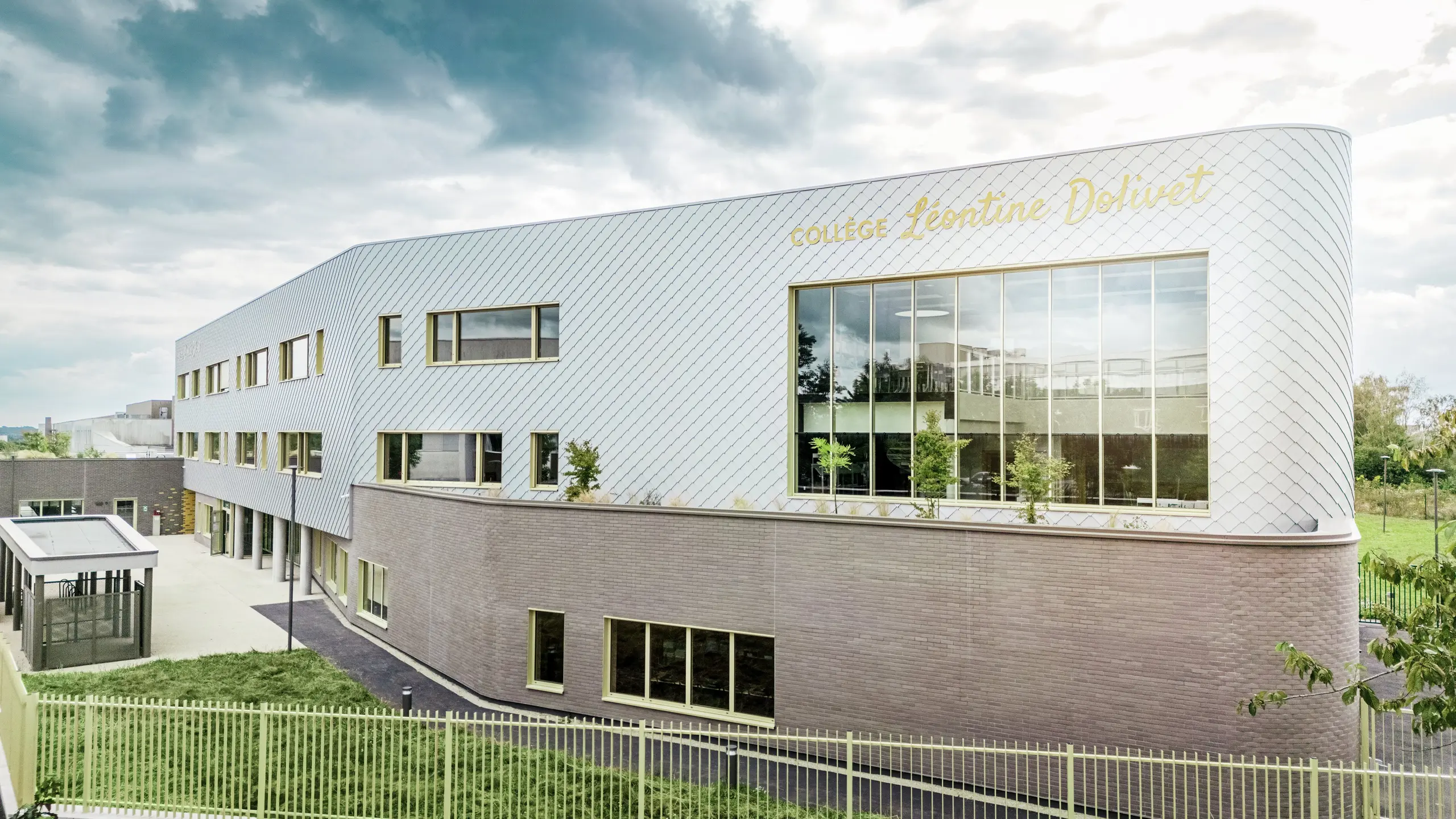 Bočni pogled na Collège Léontine Dolivet u Cesson-Sévignéu s modernom aluminijskom fasadom od PREFA zidnih rombova u srebrno-metalnoj boji. Dijagonalna struktura stvara dinamičan izgled, dok velike staklene površine i žuti prozorski okviri dodaju zanimljive naglaske. U prvom planu vidljive su zelene površine i ograda koja uokviruje zgradu. Dugotrajno PREFA rješenje fasade spaja eleganciju, otpornost na vremenske uvjete i održivost – arhitektonski privlačan objekt s jasnim linijama i promjenjivim metalom.