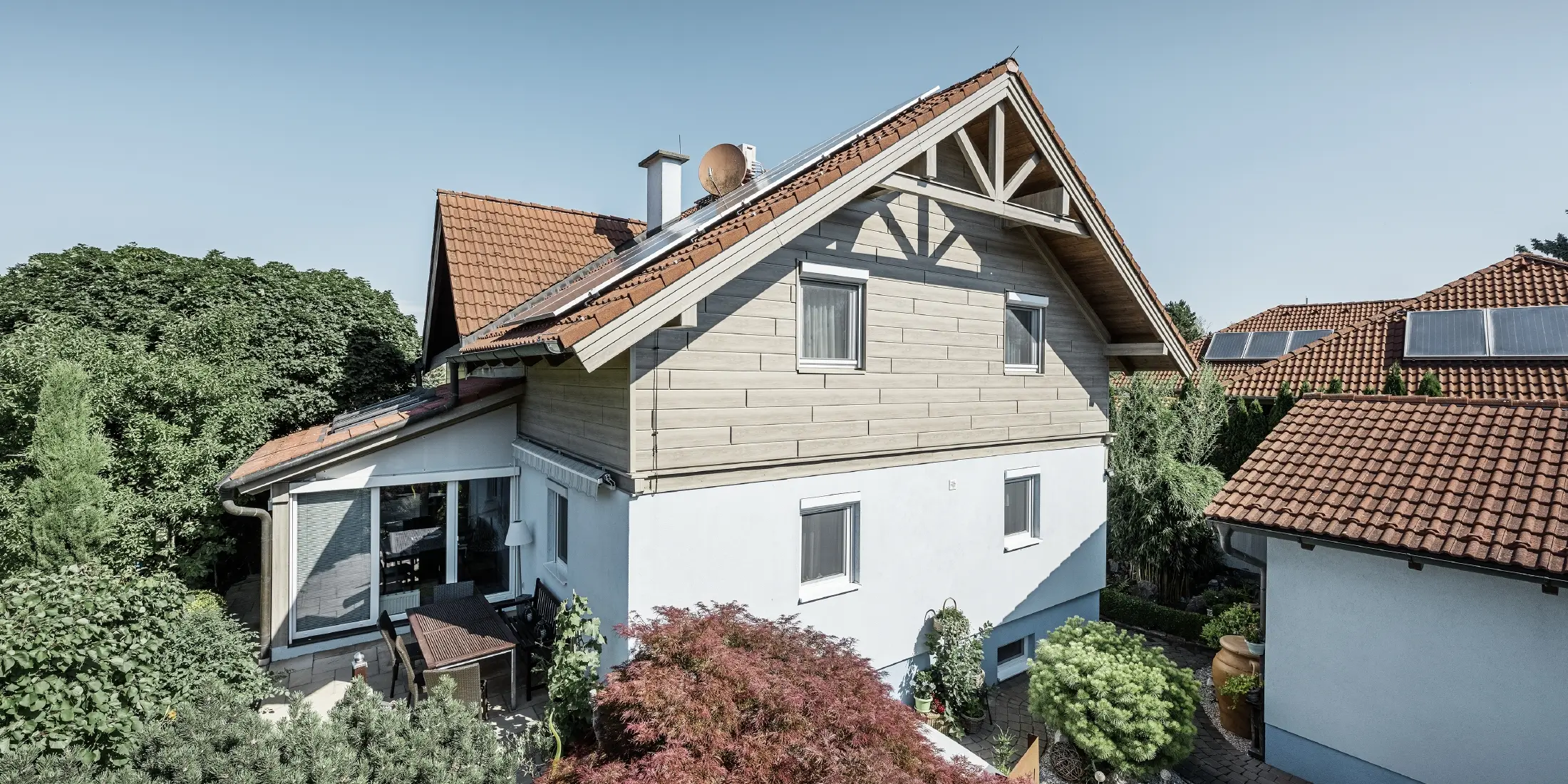 Samostojeća kuća s krovom na dvije vode i crvenim krovnim pokrovom, u kombinaciji s modernim dizajnom pročelja. Gornji zabat obložen je bež-sivim aluminijskim fasadnim panelima tvrtke PREFA, dok je prizemlje obloženo bijelim žbukanim fasadom. Na krovu su postavljeni solarni paneli. Kuća je smještena u zelenom vrtu s terasom, vrtnim namještajem i bujnim nasadima, okružena drugim stambenim zgradama slične krovne arhitekture.