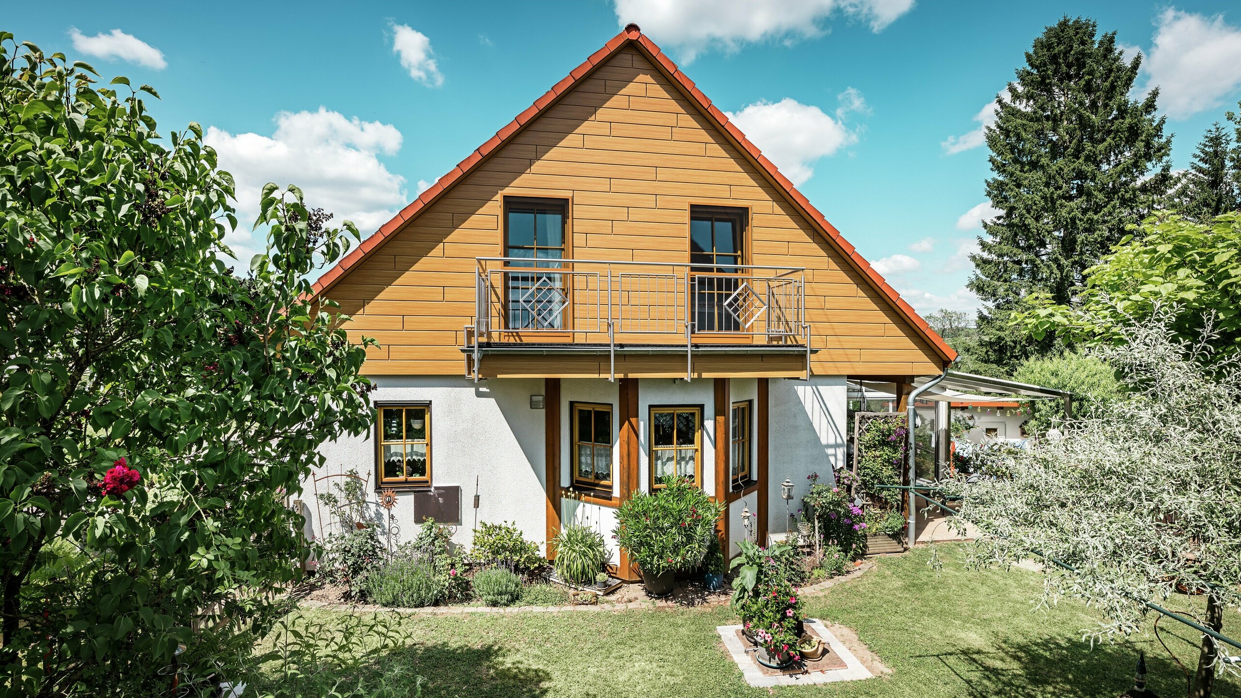 Na ovoj fotografiji, obnovljeni zabat samostojeće kuće u Blieskastelu blista u toploj prirodnoj hrastovoj nijansi zahvaljujući PREFA oblogama. Visokokvalitetna aluminijska fasada, sa svojim realističnim završnim obradom u drvenom efektu, stvara upečatljiv vizualni element i impresivno se uklapa s crvenim krovom od crijepa i bujnim vrtom. Okružen cvijećem, grmljem i bujnim zelenilom, kuća izgleda privlačno i dobro održavano. PREFA površina kombinira prirodan izgled s funkcionalnim prednostima kao što su visoka otpornost na vremenske uvjete, stabilnost i gotovo održavanje – estetski vrhunac s održivom dodanom vrijednošću.