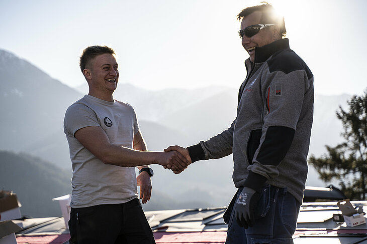 Na kosom krovu kuće stoje PREFA trener i limar koji se rukuju. Obojica nose profesionalnu radnu odjeću i zaštitnu opremu. U pozadini se vidi nisko sunce i pakirne kutije PREFA aluminijskih proizvoda. Ova scena simbolizira uspješnu suradnju i stručnu razmjenu s kvalitetom rukujućeg dogovora. PREFA trener podržava limara svojim znanjem o obradi, tehnologiji i proizvodnim sustavima. Slika prikazuje blisku podršku koju PREFA pruža obrtnicima – od edukacije do profesionalne primjene na gradilištu.