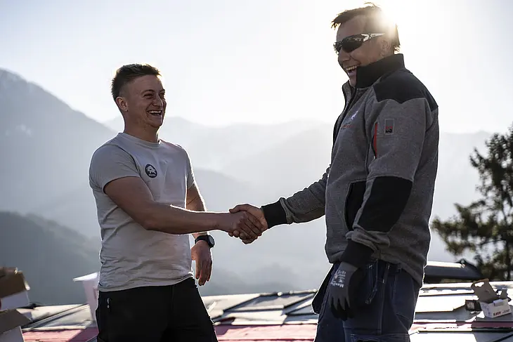 Na kosom krovu kuće stoje PREFA trener i limar koji se rukuju. Obojica nose profesionalnu radnu odjeću i zaštitnu opremu. U pozadini se vidi nisko sunce i pakirne kutije PREFA aluminijskih proizvoda. Ova scena simbolizira uspješnu suradnju i stručnu razmjenu s kvalitetom rukujućeg dogovora. PREFA trener podržava limara svojim znanjem o obradi, tehnologiji i proizvodnim sustavima. Slika prikazuje blisku podršku koju PREFA pruža obrtnicima – od edukacije do profesionalne primjene na gradilištu.