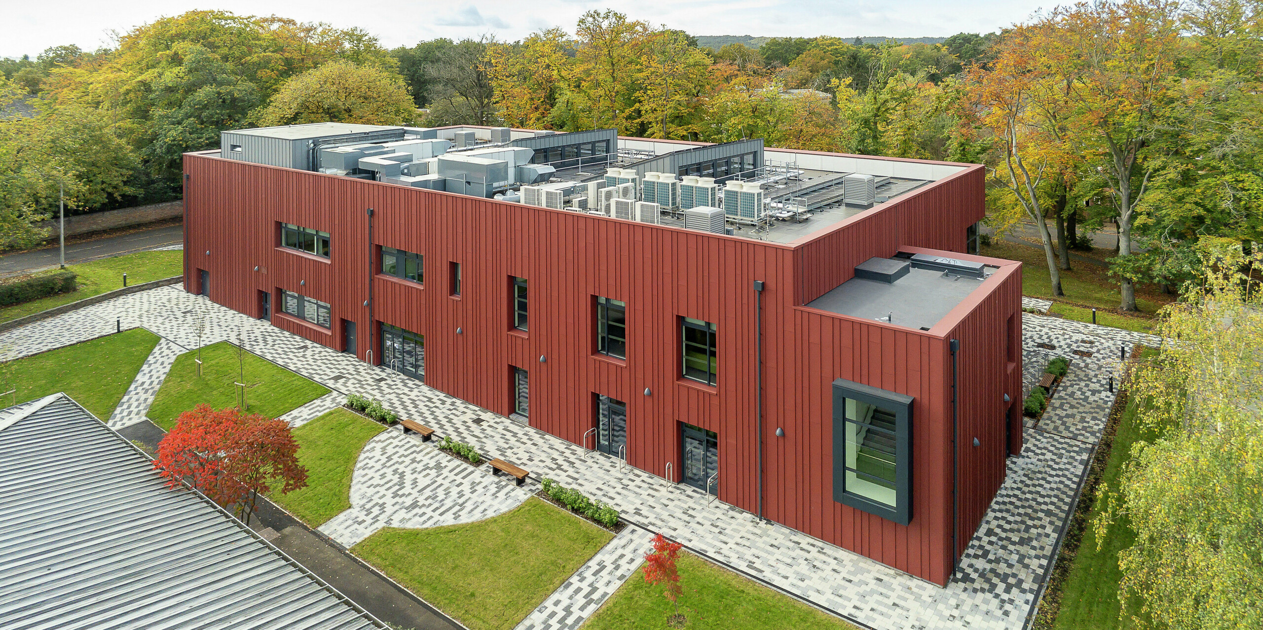 Architektur im Einklang mit der Natur: Das Soldiers' Centre in Pirbright aus der Vogelperspektive. Die markante Stehfalzfassade aus PREFALZ in P.10 Oxydrot fügt sich harmonisch in die grüne Umgebung ein. Die auskragenden Fensterrahmen in P.10 Anthrazit setzen moderne Akzente. Die strukturierte Außenanlage mit gepflasterten Wegen, Grünflächen und Sitzgelegenheiten schafft eine einladende Atmosphäre. Auf dem Flachdach sind technische Installationen integriert, die zur modernen Gebäudetechnik beitragen.