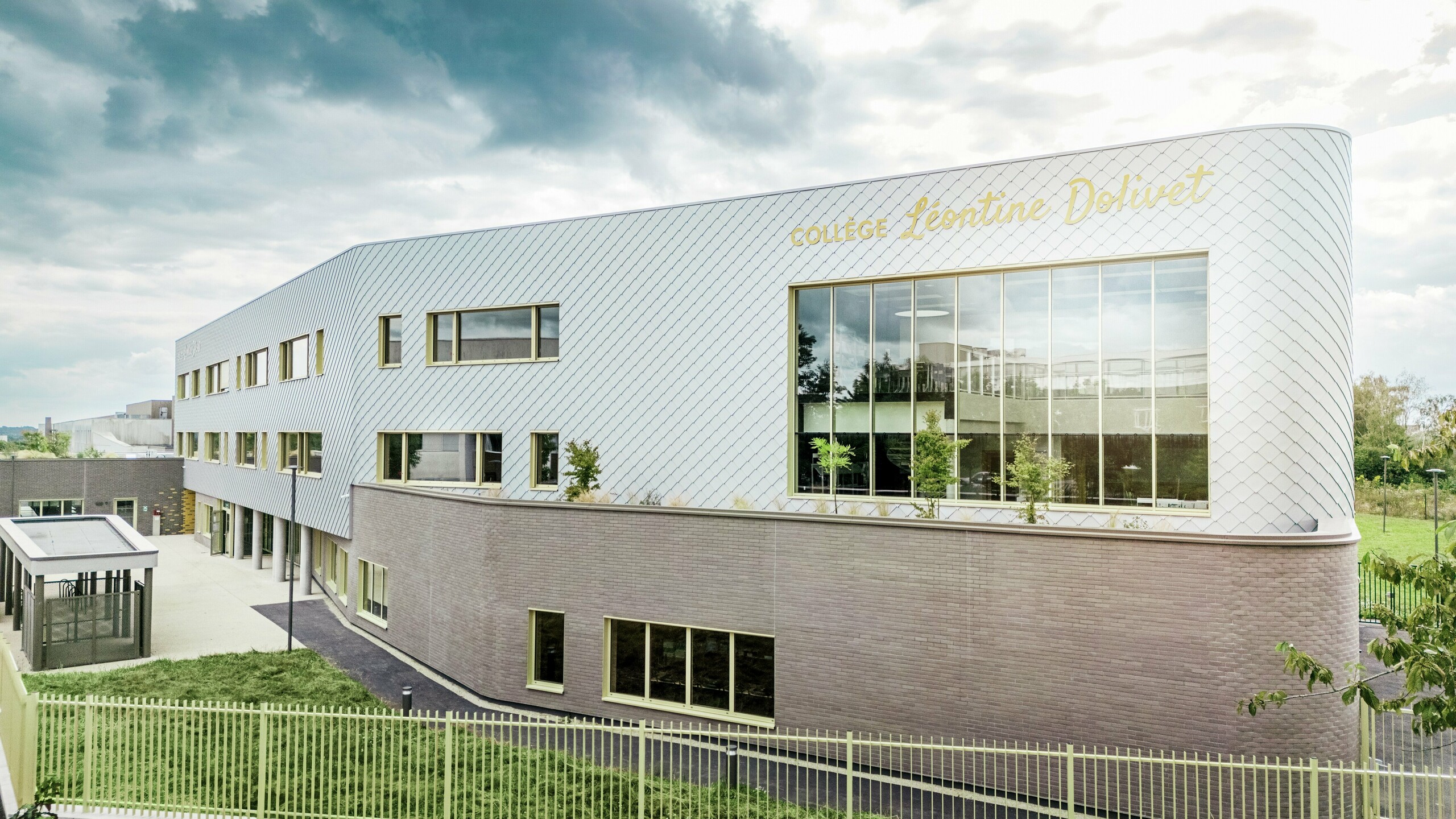 Bočni pogled na Collège Léontine Dolivet u Cesson-Sévignéu s modernom aluminijskom fasadom od PREFA zidnih rombova u srebrno-metalnoj boji. Dijagonalna struktura stvara dinamičan izgled, dok velike staklene površine i žuti prozorski okviri dodaju zanimljive naglaske. U prvom planu vidljive su zelene površine i ograda koja uokviruje zgradu. Dugotrajno PREFA rješenje fasade spaja eleganciju, otpornost na vremenske uvjete i održivost – arhitektonski privlačan objekt s jasnim linijama i promjenjivim metalom.