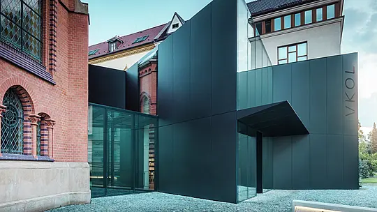 Slika prikazuje modernu nadogradnju knjižnice koja se elegantno nadovezuje na povijesnu Crvenu crkvu u Olomoucu. Zapanjujući novi objekt impresionira fasadom od crnih PREFABOND aluminijsko-kompozitnih ploča, oblikovanih u čistim, geometrijskim linijama i izrađenih s preciznom pažnjom na detalje. Stakleni zidovi zgrade omogućuju transparentan pogled u unutrašnjost, koja se skladno uklapa u povijesno okruženje. Igra modernih materijala i klasičnih arhitektonskih elemenata stvara zanimljivu vizualnu povezanost između starog i novog. Inovativna primjena PREFABOND ploča osigurava ne samo moderan izgled, već i dugovječnost i otpornost, pripremajući zgradu za budućnost.