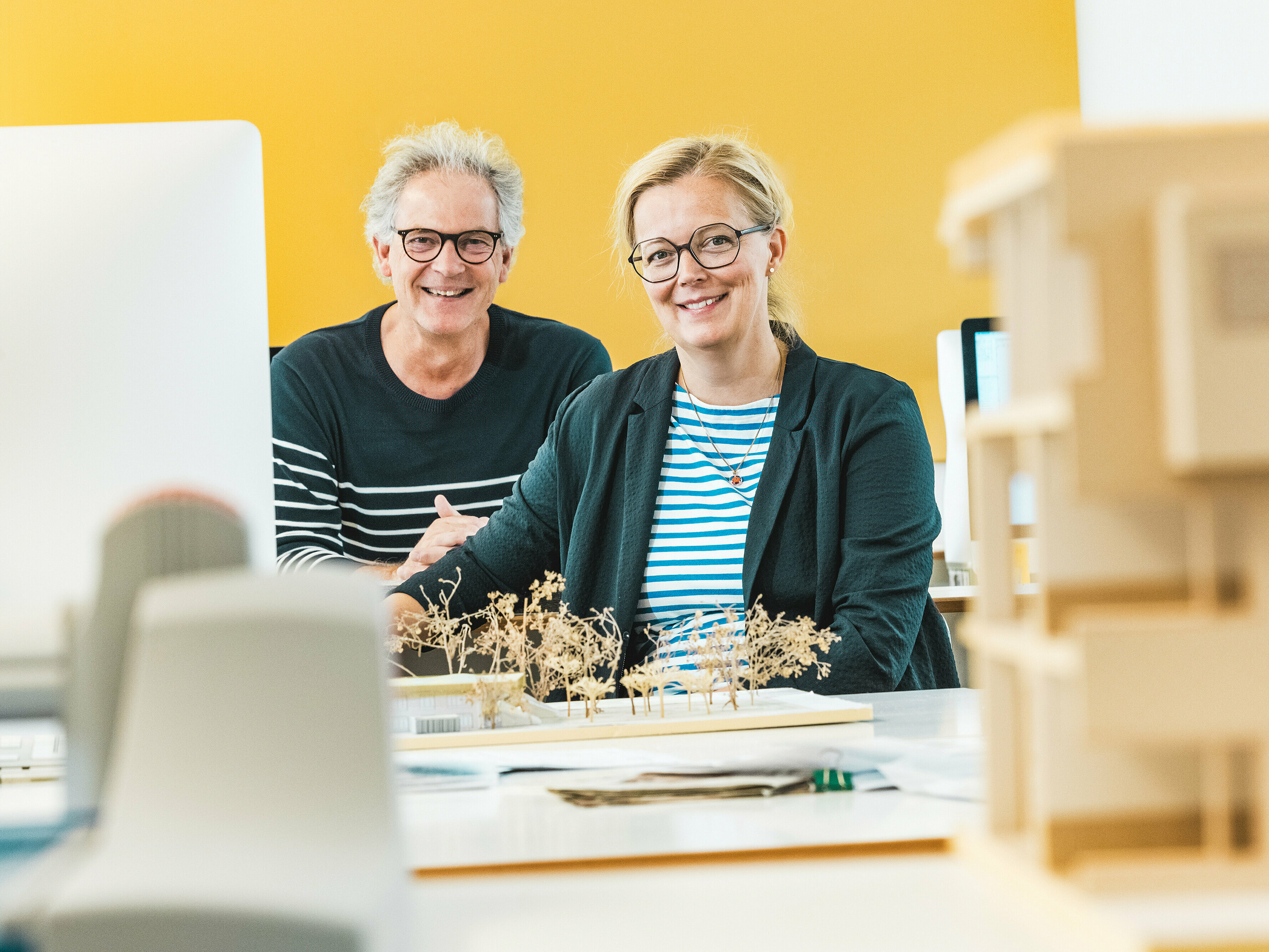 Arhitekti Claudia i Theo Härtner sjede blistajući ispred arhitektonskog modela. Ovaj arhitektonski par realizira dječji vrtić u Alzenauu, budućnosno orijentiranu hibridnu zgradu s fasadom od PREFA aluminija. U prvom planu modela vidljive su minijaturne drvene figure koje odražavaju održivi koncept cijelog projekta. Dječji vrtić u Alzenauu spaja inovativnu arhitekturu s odgovornim urbanim razvojem i pokazuje svestranost PREFA zidnih rombova 44 × 44 u brončanoj boji. Dugotrajna, jednostavna za održavanje i u potpunosti reciklabilna aluminijska fasada vizualno povezuje stambeni i vrtićki dio u skladnu cjelinu. Slika hvata kreativnu energiju procesa planiranja i naglašava estetske i ekološke prednosti PREFA proizvoda.